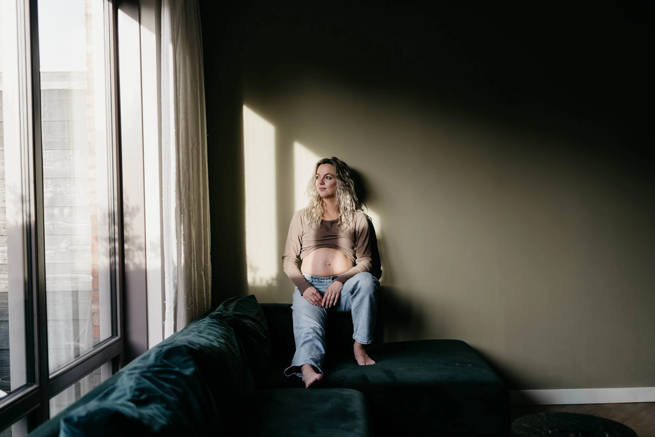 A woman with curly blonde hair sitting on the arm of a dark green couch in a room with green wall, sunlight streaming through large windows with white curtains.