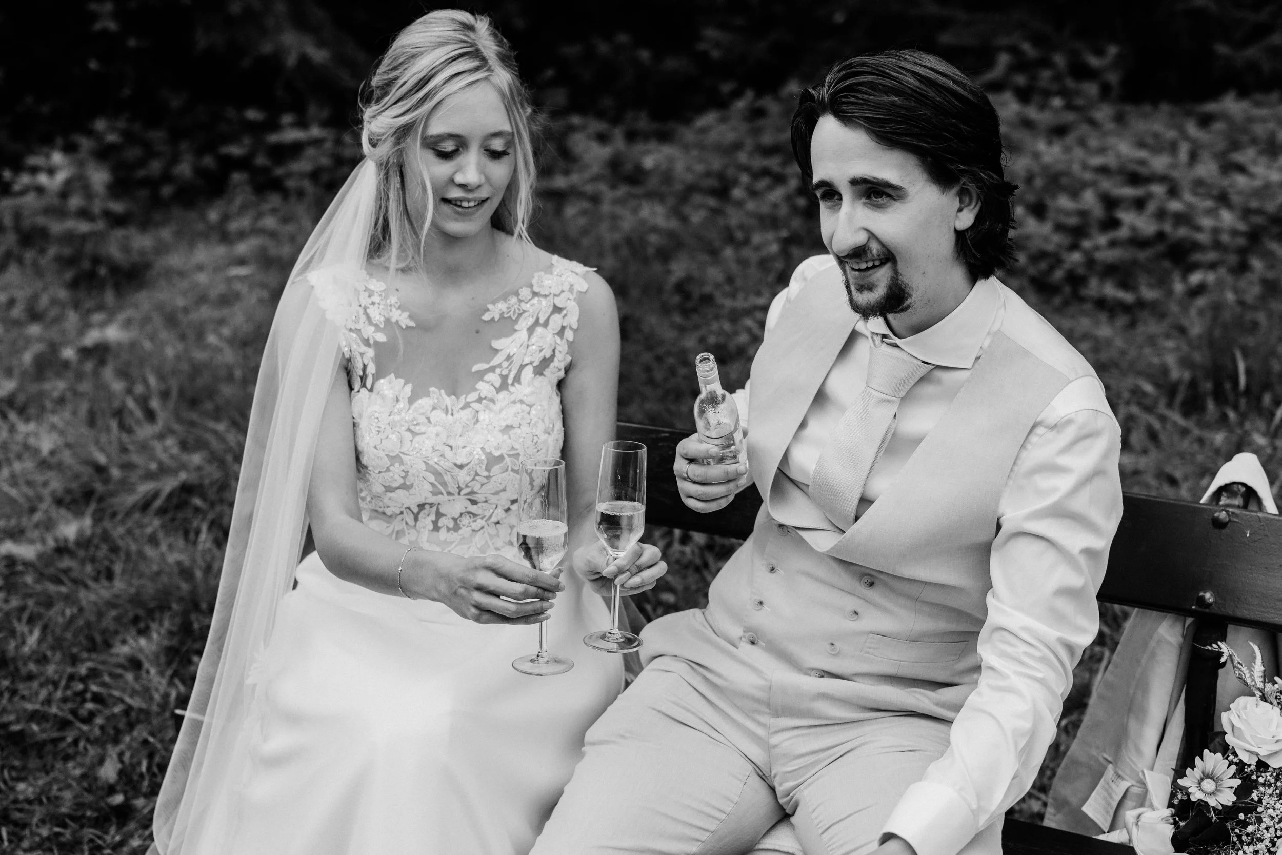 A bride and groom sitting on a park bench, holding champagne glasses, smiling and enjoying each other's company. The bride is in a lace wedding dress with a veil, and the groom is in a light-colored suit with a tie, during a wedding celebration outdo