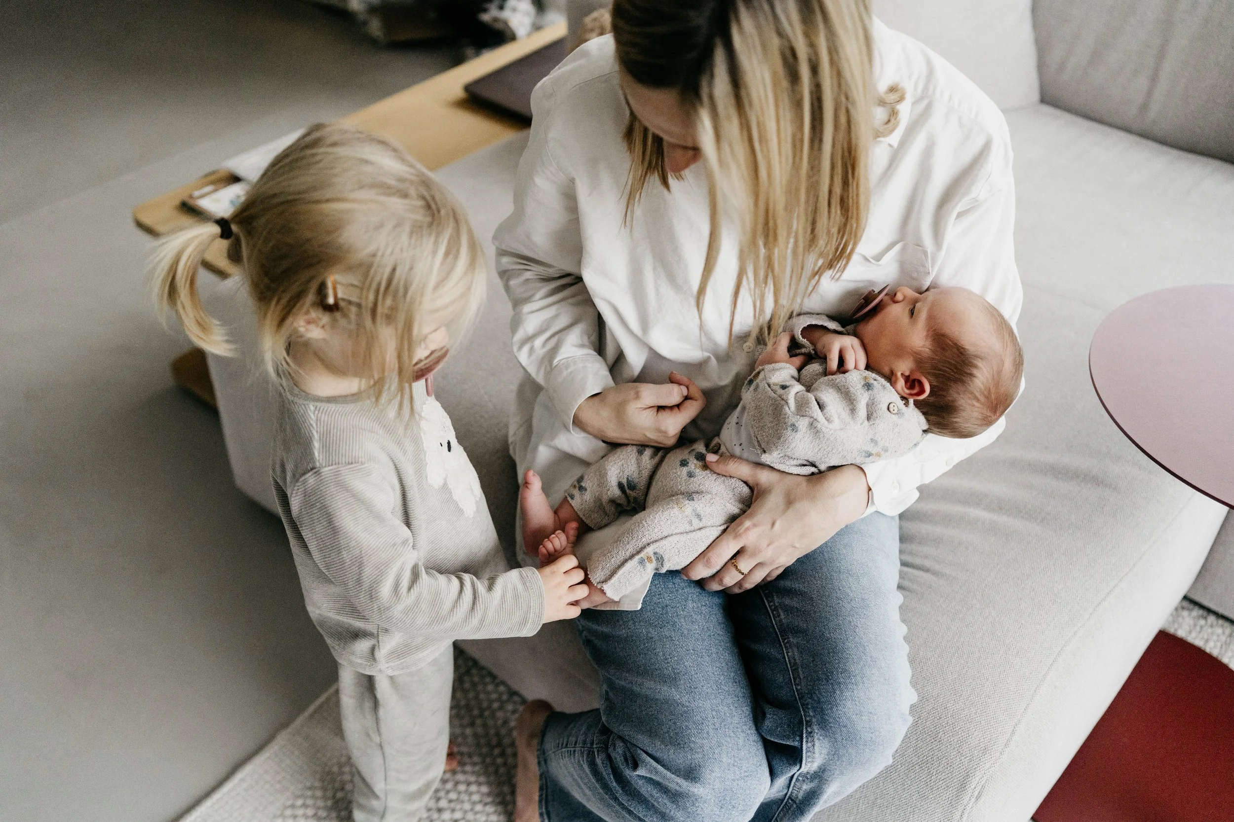 A woman holding a sleeping baby in her arms while a young girl touches the baby's feet in a cozy living room.