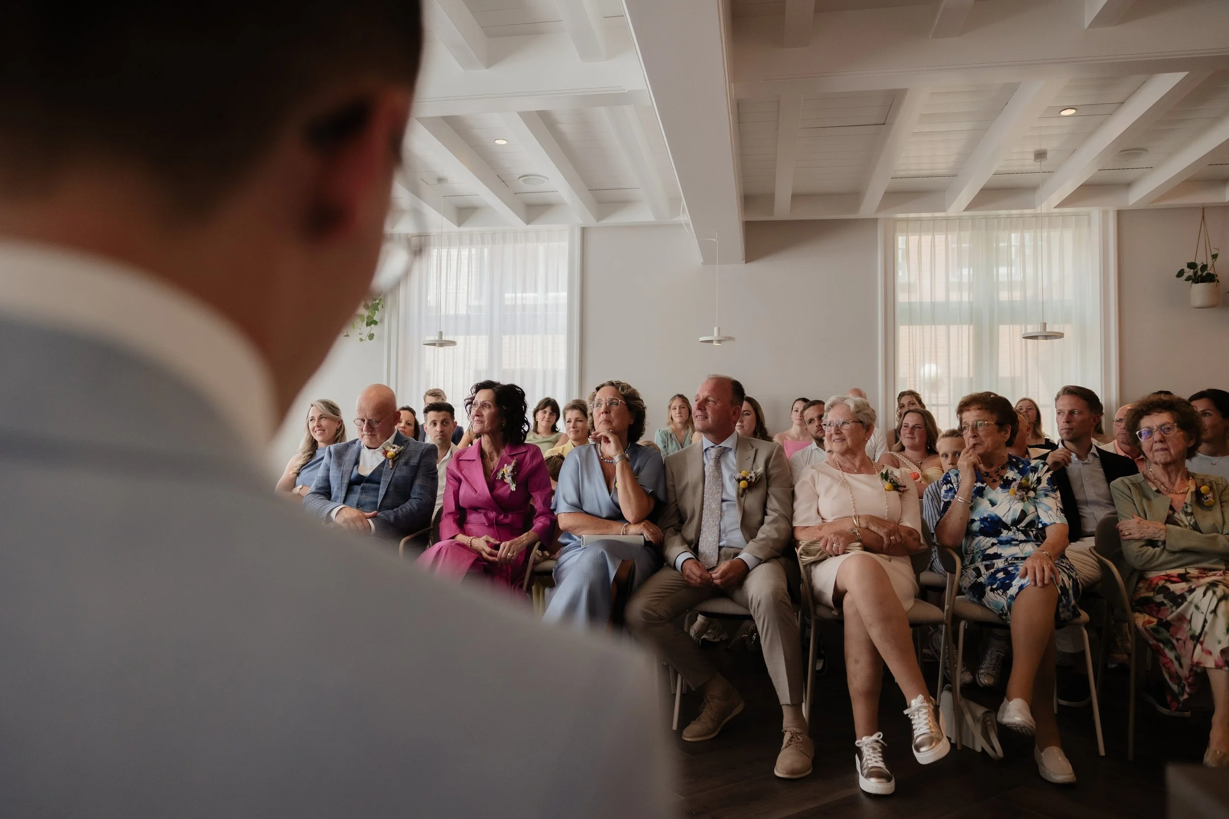 The image shows a guest attending a wedding ceremony, seated among other guests in a well-lit room with white walls and large windows. The focus is on the guest, wearing a suit, with their back to the camera, facing the seated guests.