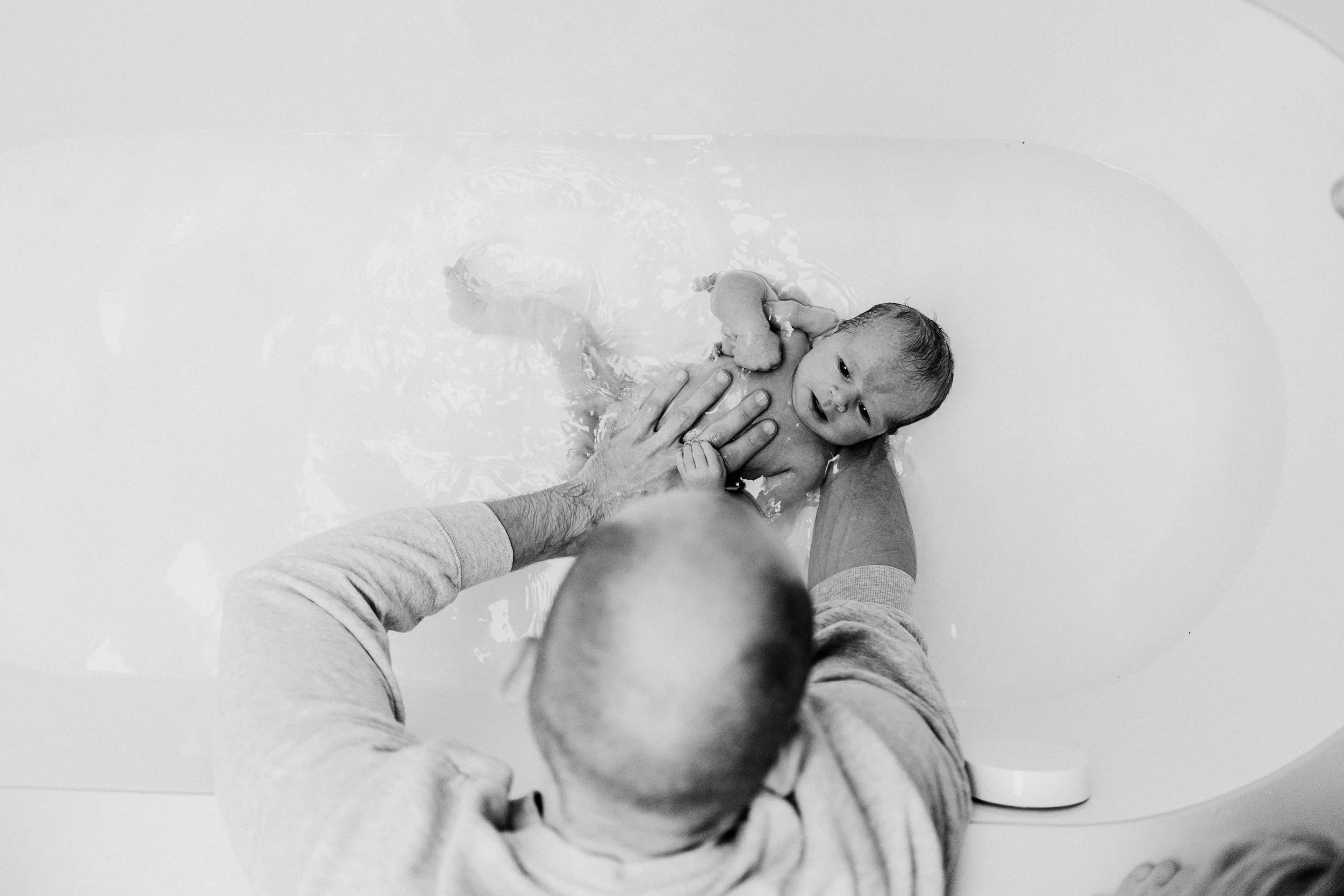 A man helping a small baby in a bathtub, with the baby smiling and the man's arm visible from above.