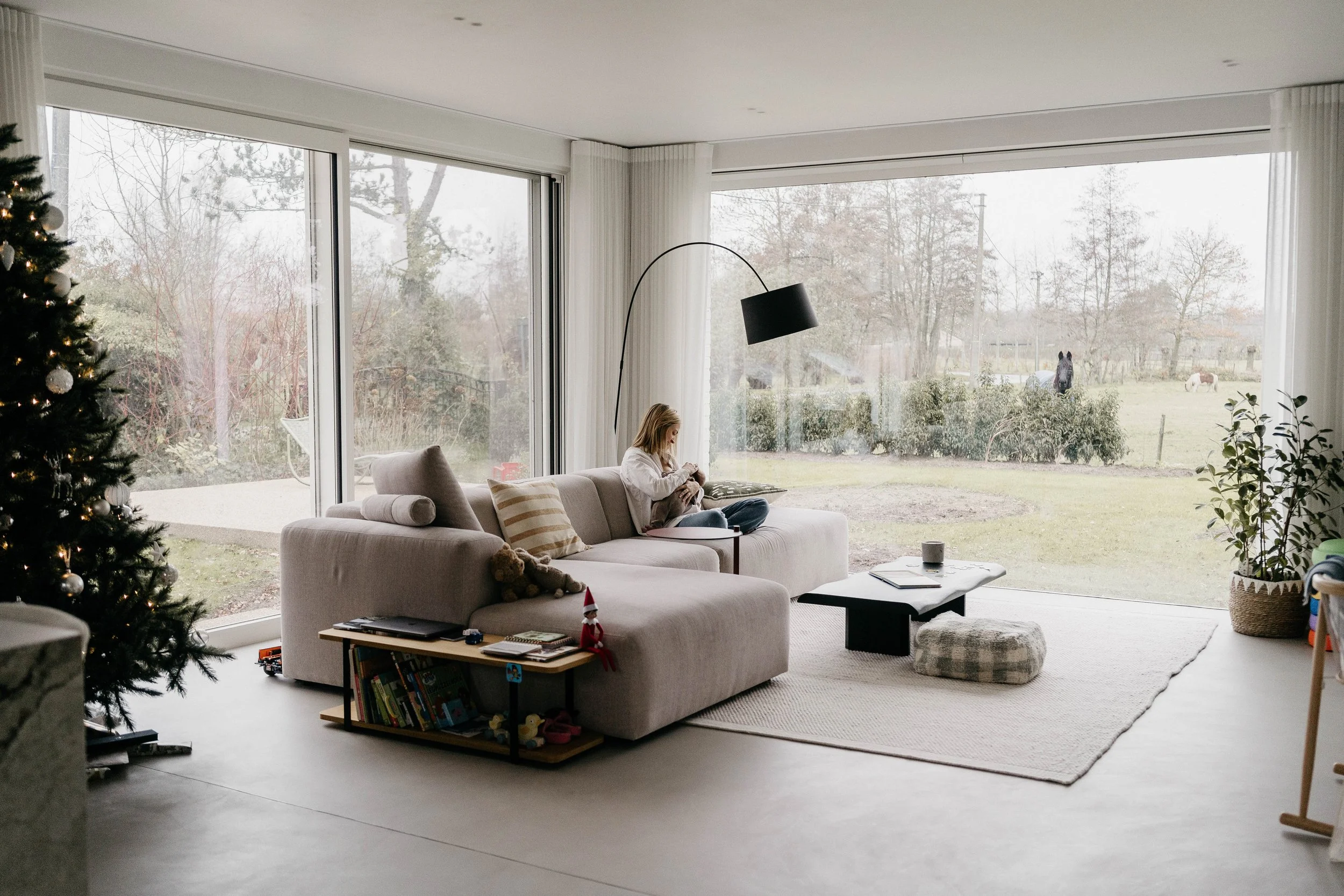 A woman sitting on a beige couch in a modern living room with large windows overlooking a backyard with trees and horses.