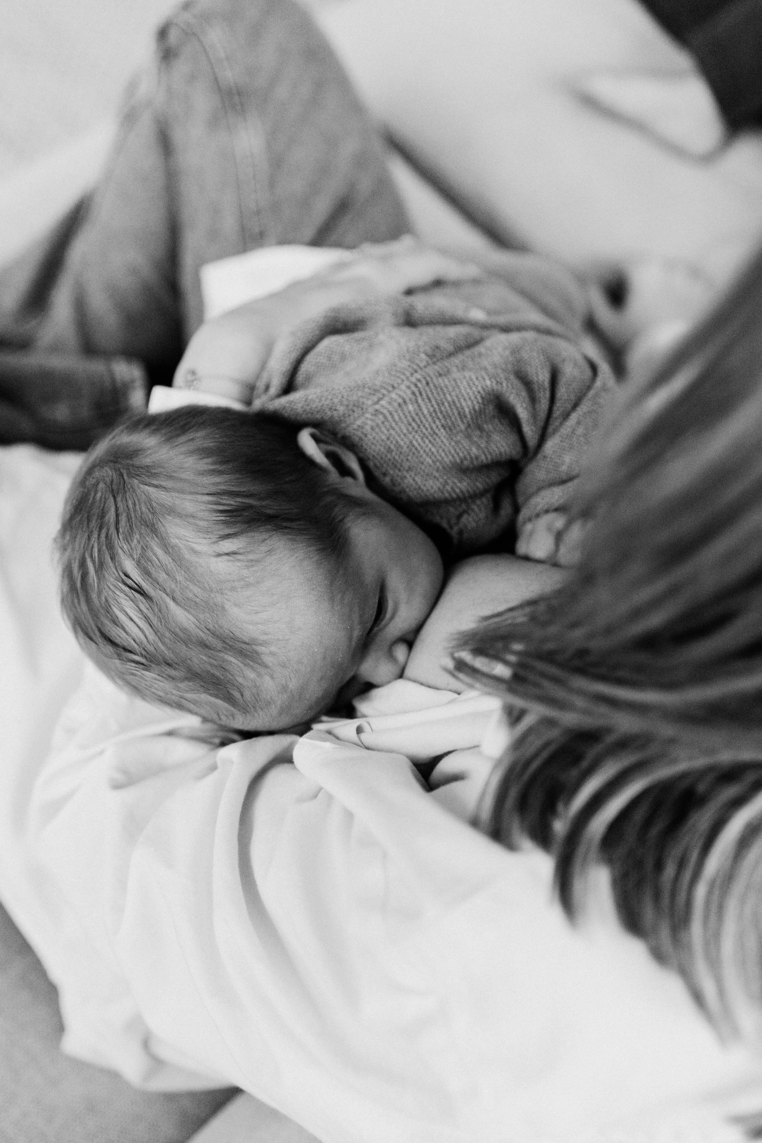 A young child lying on a bed, resting on their stomach, with a blanket partially covering them, while a woman with long hair is close by, possibly comforting or watching over the child.