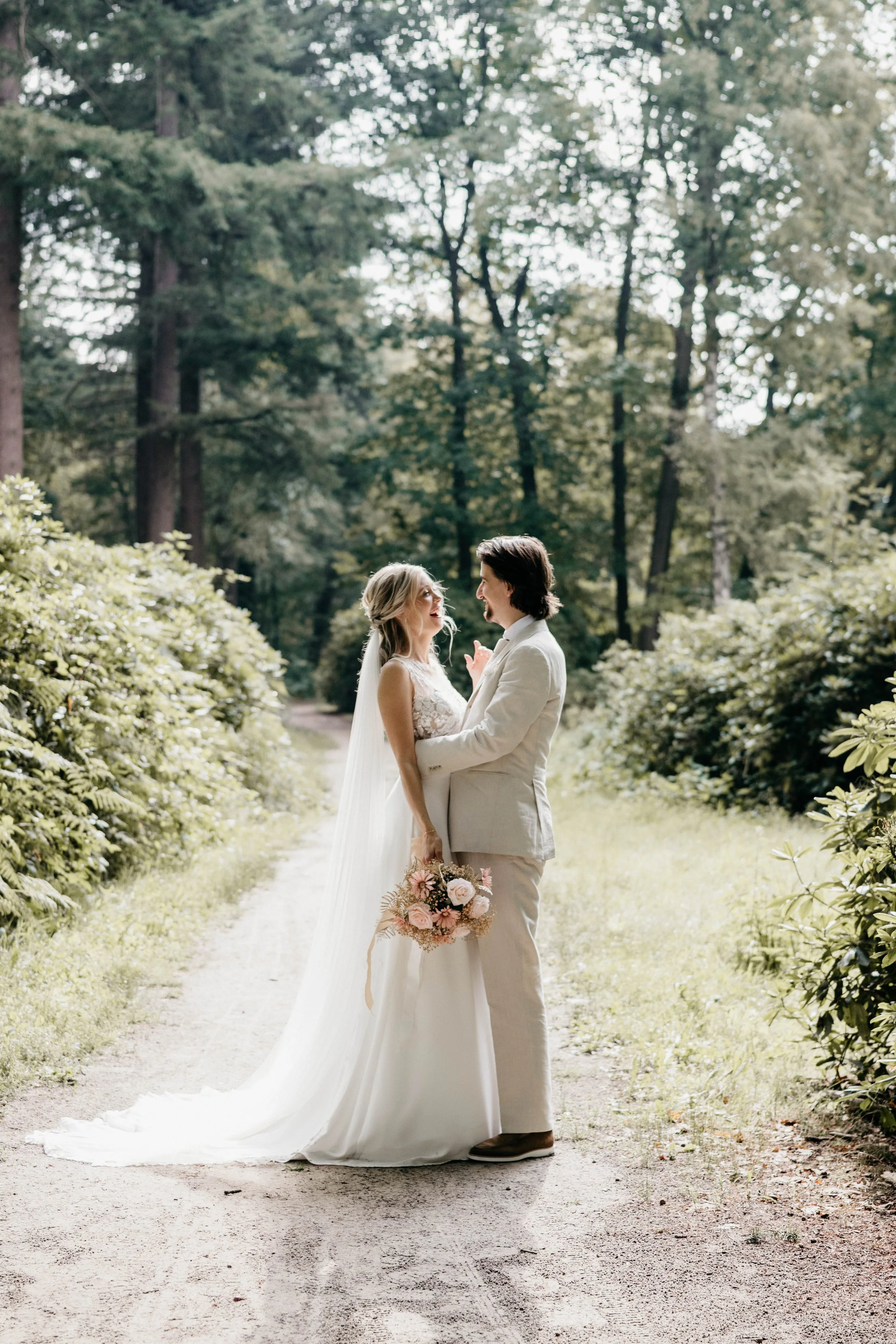 A bride and groom standing on a forest trail, holding hands and gazing at each other, surrounded by lush greenery, with the bride carrying a bouquet of pink and white flowers.