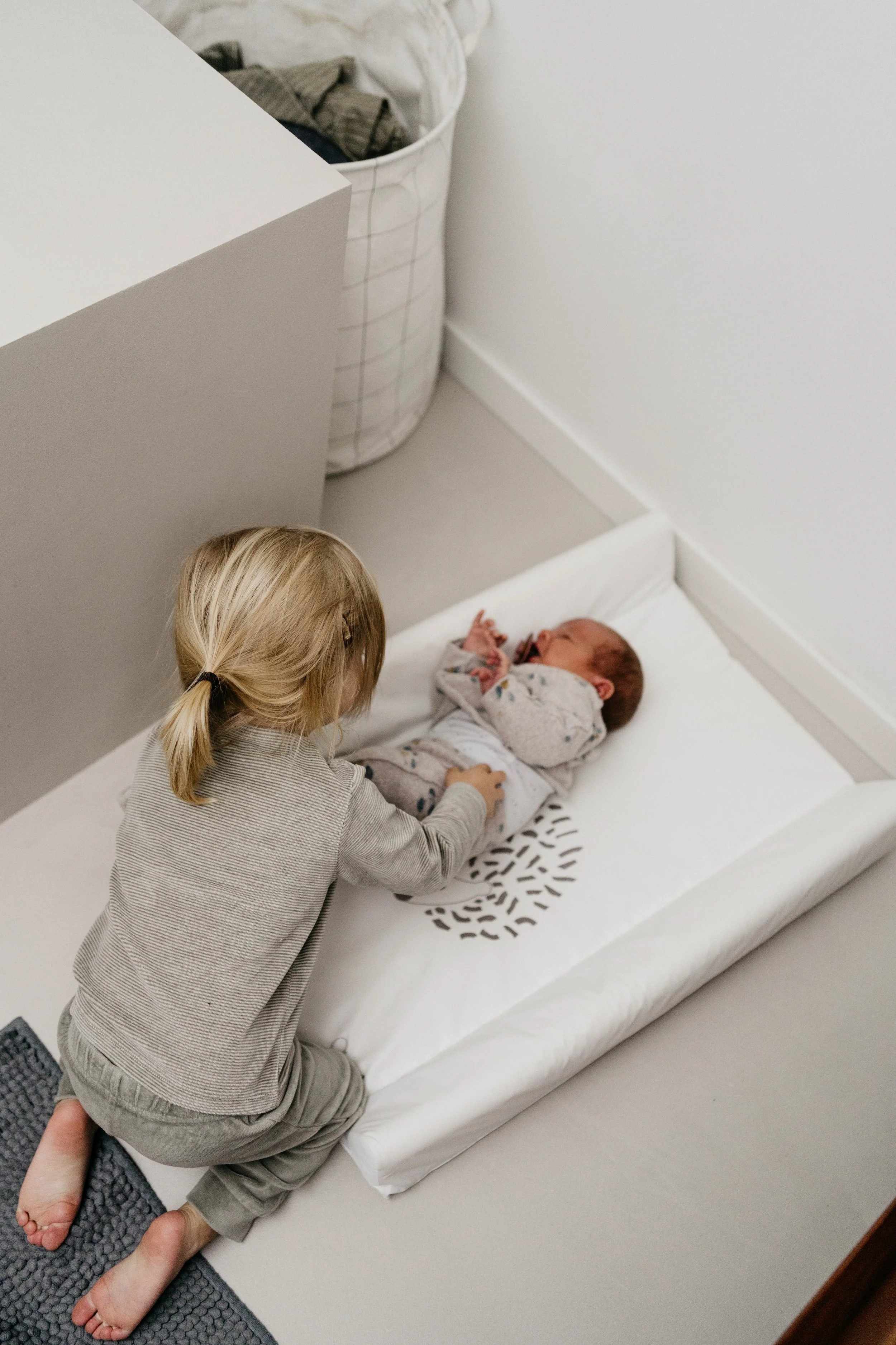 A young girl with blonde hair in a ponytail kneeling on a white changing pad, looking at and interacting with a newborn baby lying on the pad.