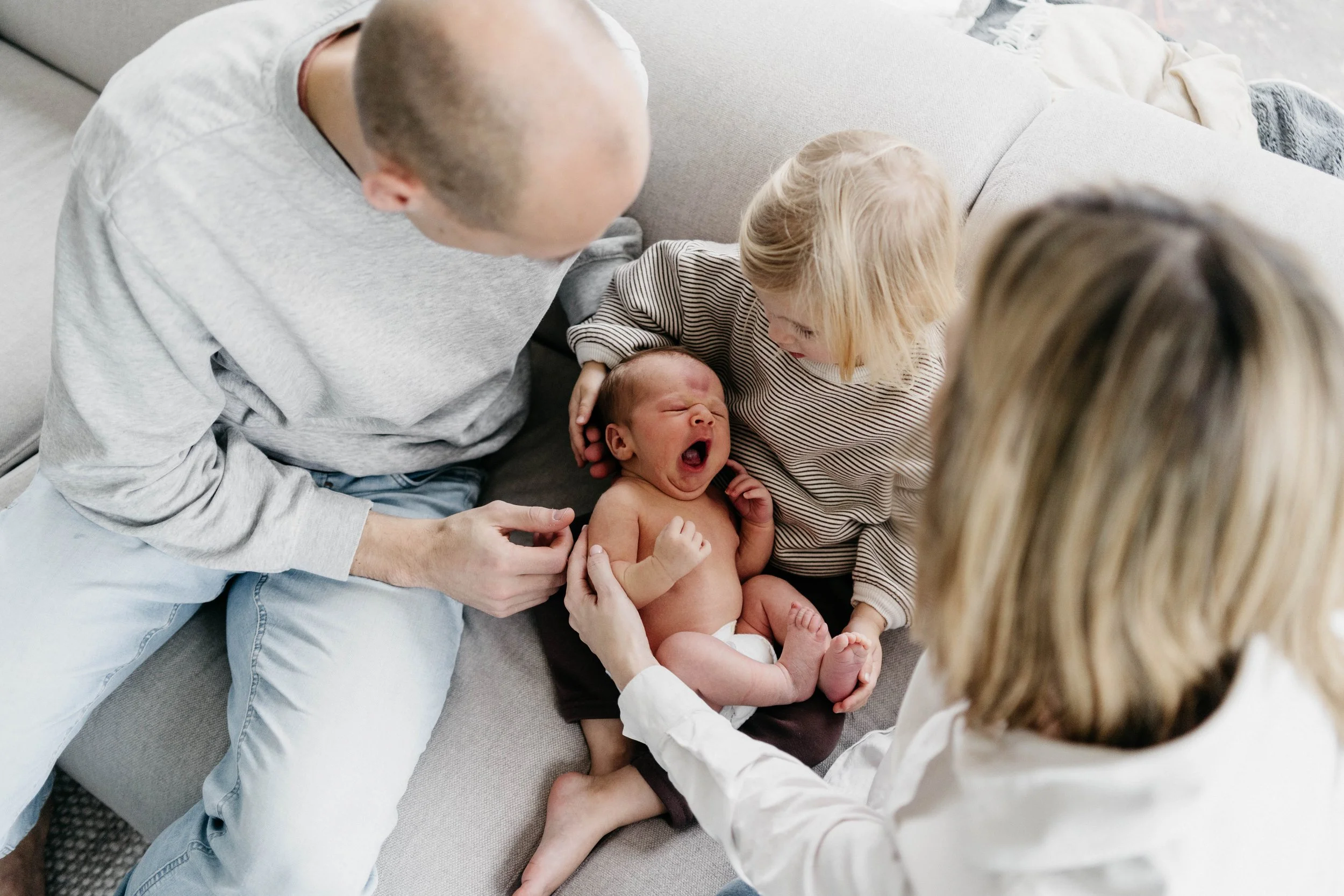 Infant yawning while being held by a young girl, with a man and woman nearby, on a gray couch in a living room.