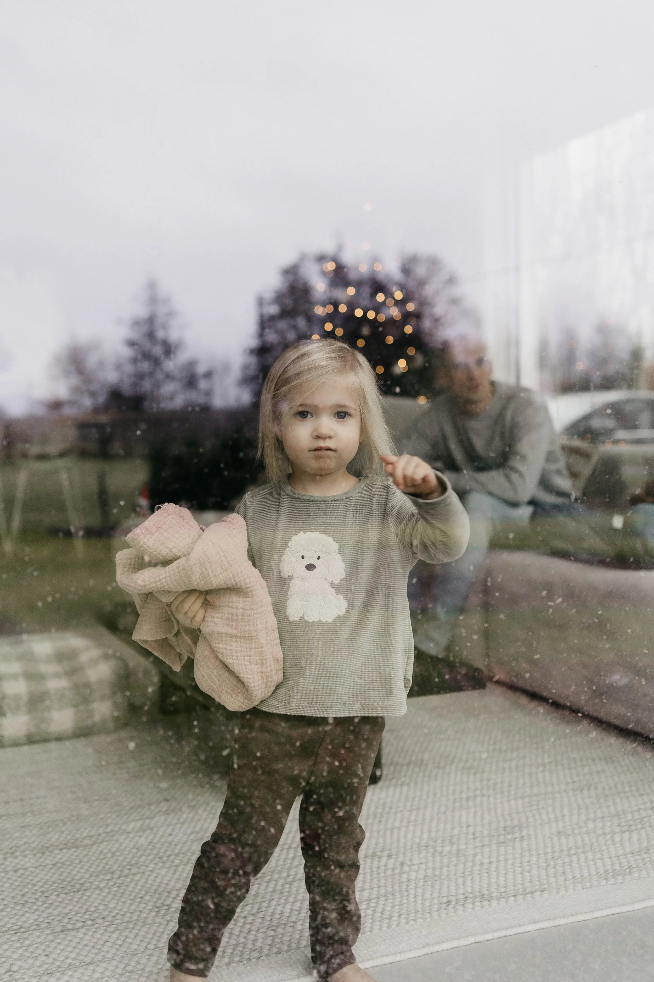 A young girl with blonde hair and blue eyes, standing behind a glass door, holding a blanket, with a reflection of a woman and a decorated Christmas tree visible through the glass on a cloudy day.