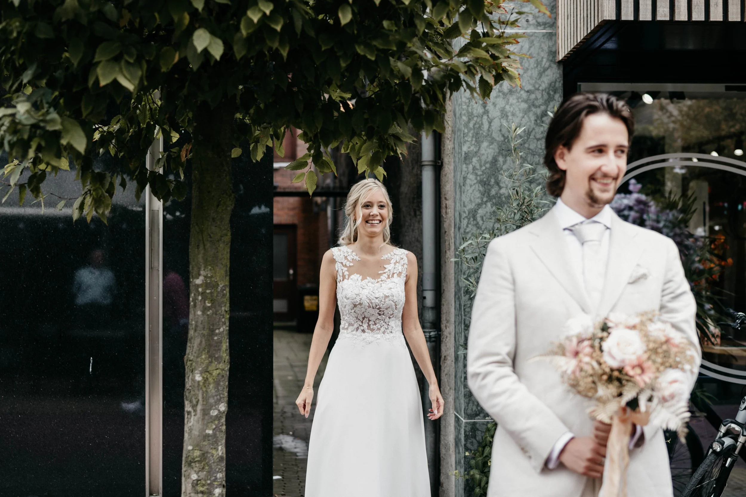A bride and groom outside, with the bride smiling and the groom holding a bouquet of flowers, on a city street.