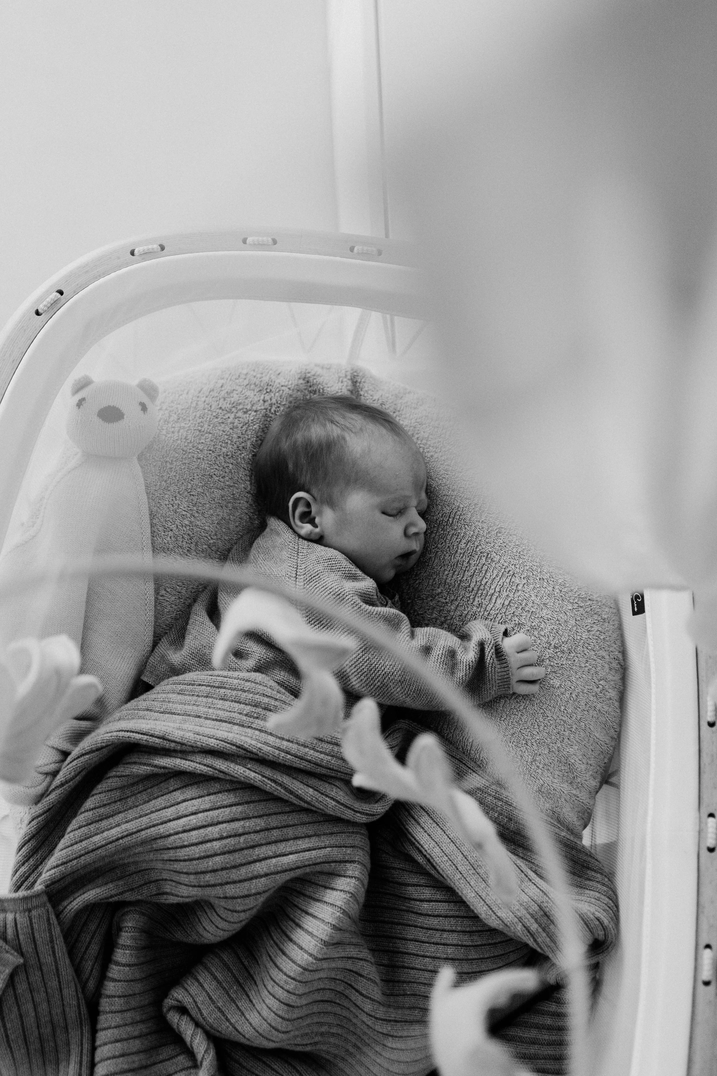A sleeping baby lying on a cushioned surface, covered with a striped blanket, next to a stuffed animal toy in a crib.