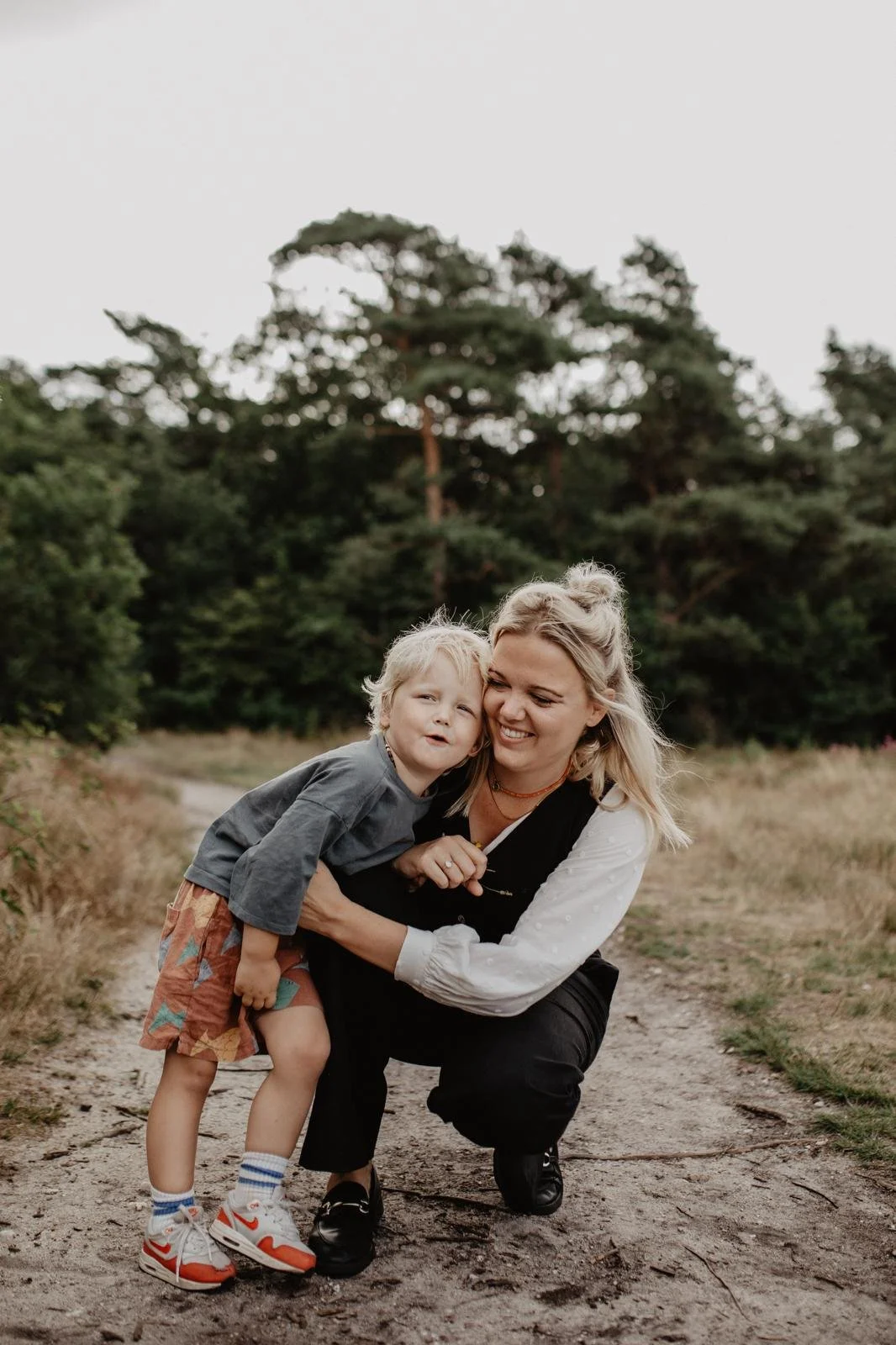 A woman and a young boy are outdoors on a dirt path surrounded by trees. The woman is squatting down, smiling and hugging the boy, who is leaning in and making a playful face. The boy is wearing a gray shirt, colorful shorts, and sneakers, and the woman is dressed in casual clothes.