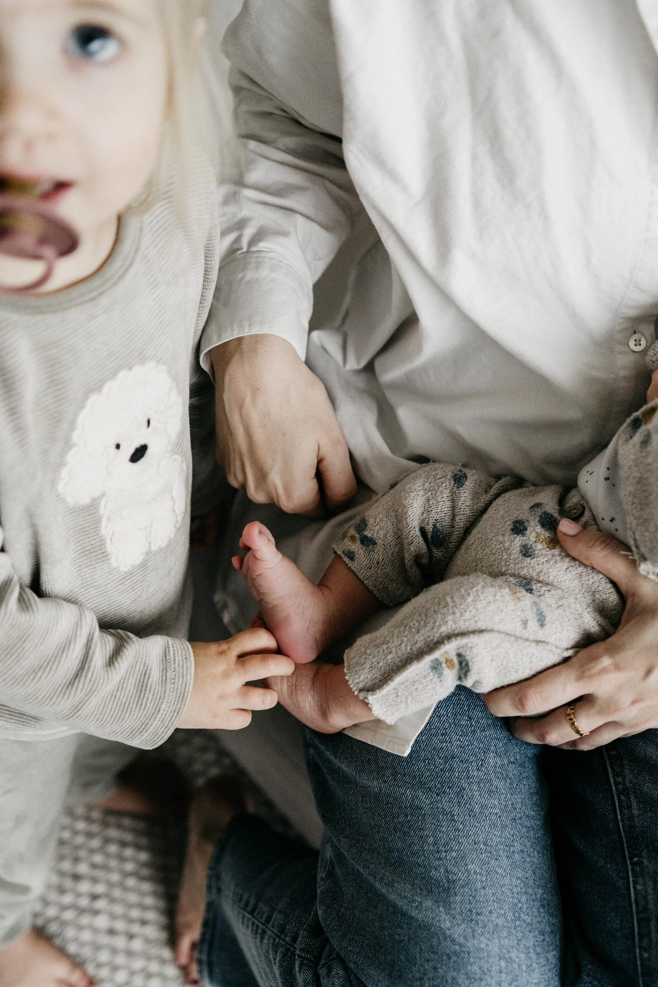 A young child with a pacifier in their mouth, reaching out to touch the foot of a baby, who is being held by an adult wearing jeans and a white shirt.