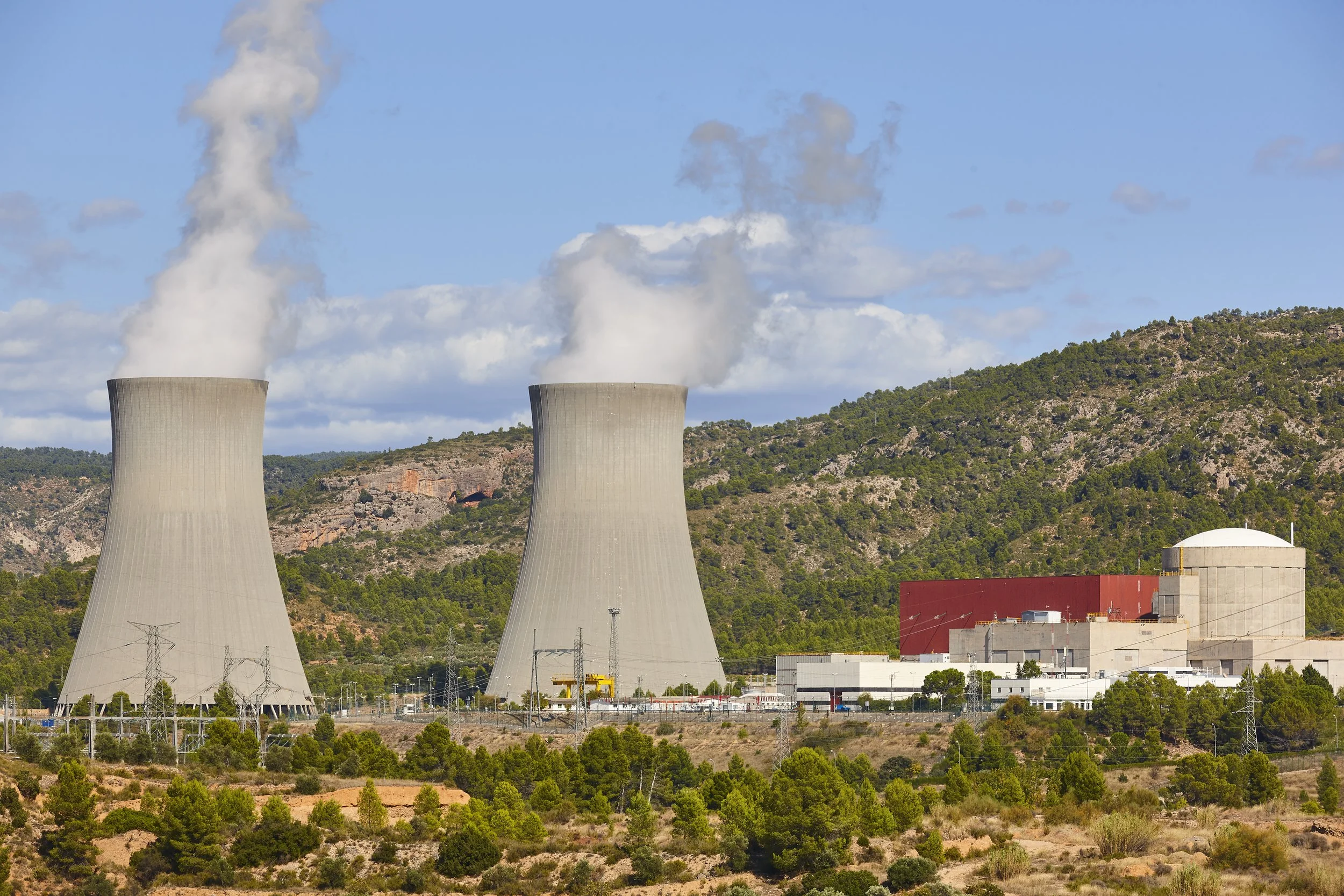 A nuclear power plant with two cooling towers emitting steam, situated in a hilly landscape with green trees and partly cloudy sky.