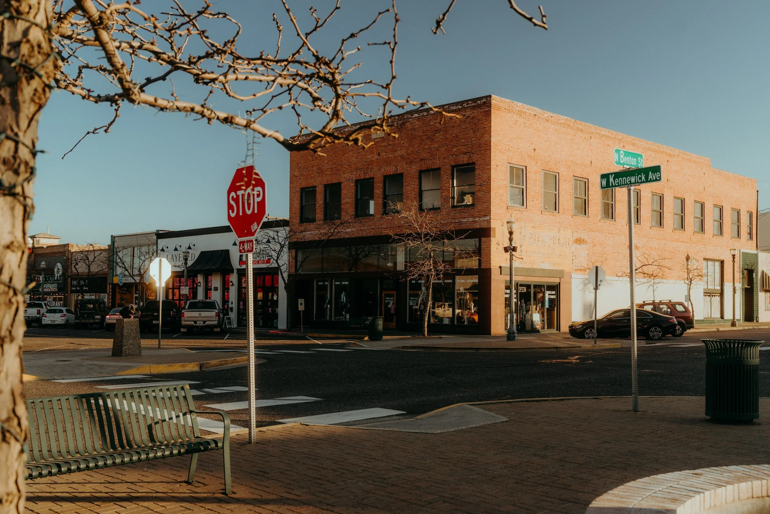 A street corner in a small town during daytime with a brick building, a stop sign, and street signs for North Benton Street and West Kennewick Avenue. There are parked cars, a bench, and leafless trees, indicating a possible winter or early spring season.