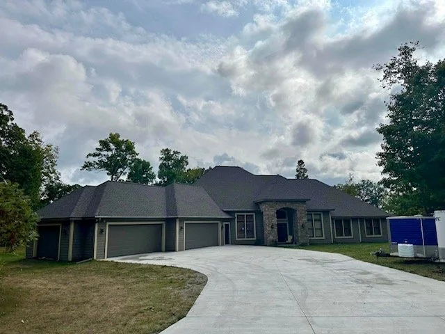 Modern house with a three-car garage, gray exterior, and a curved driveway, surrounded by trees under a cloudy sky.