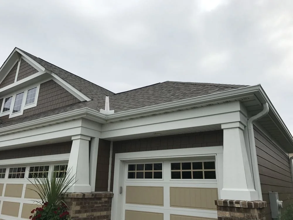 Close-up of a residential house with a brown shingle roof, beige and white garage door, and white trim, featuring a small window on the upper level and a brick and white column support at the garage.