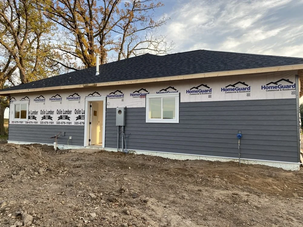 Newly constructed house with blue siding, construction materials and dirt in the foreground, and a large tree with autumn leaves behind the house.