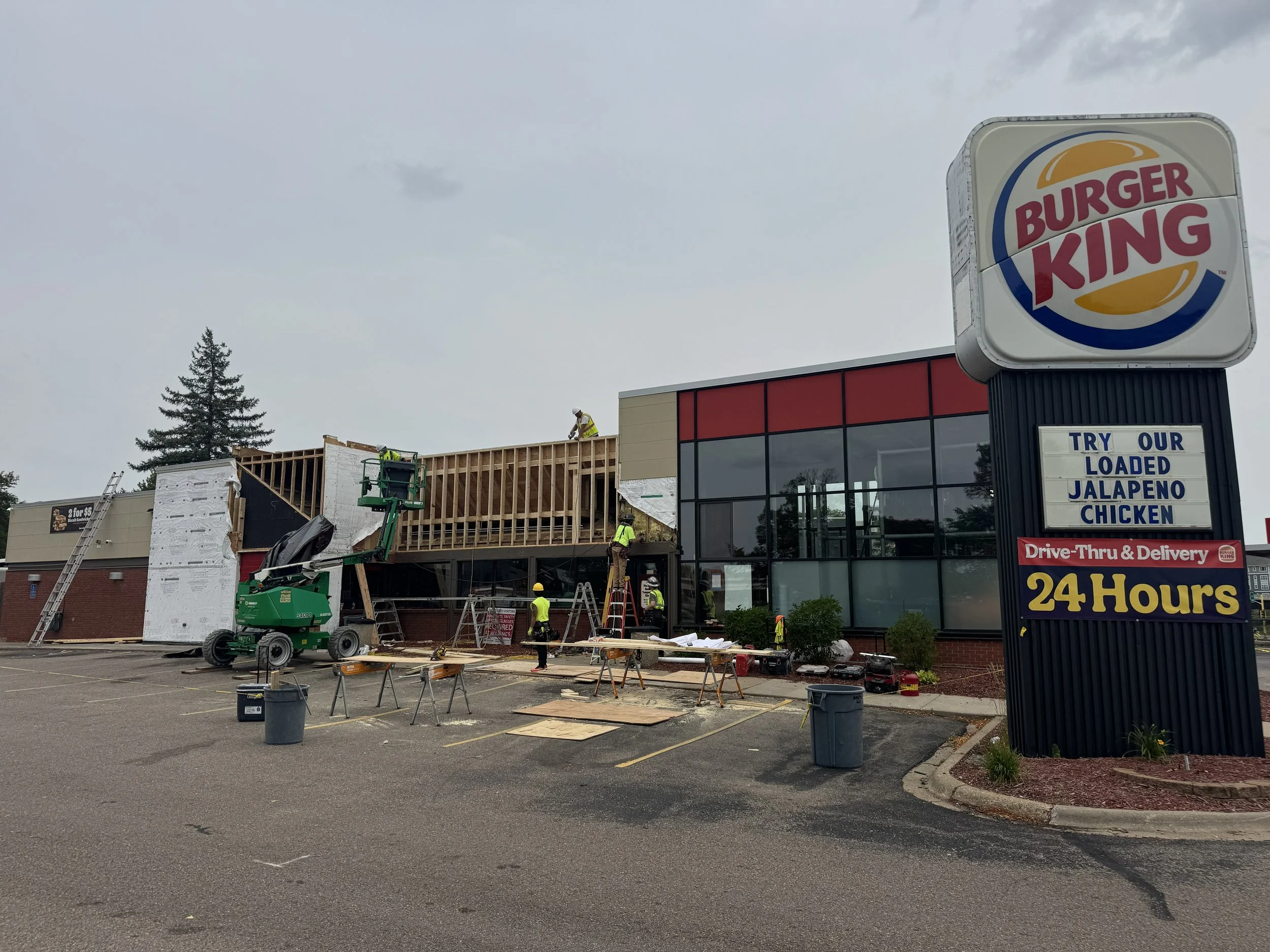 Construction workers are building an extension onto a Burger King restaurant. The partially completed structure has workers installing the roof and framing. The Burger King sign is visible, and the parking lot in the foreground is empty with some construction equipment and materials.
