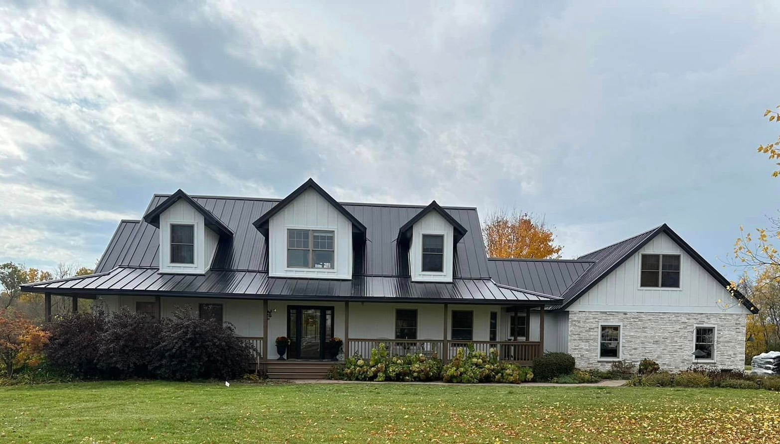 A modern house with a gabled roof and dormer windows, a wooden porch, a well-maintained lawn, autumn trees, and a partly cloudy sky.