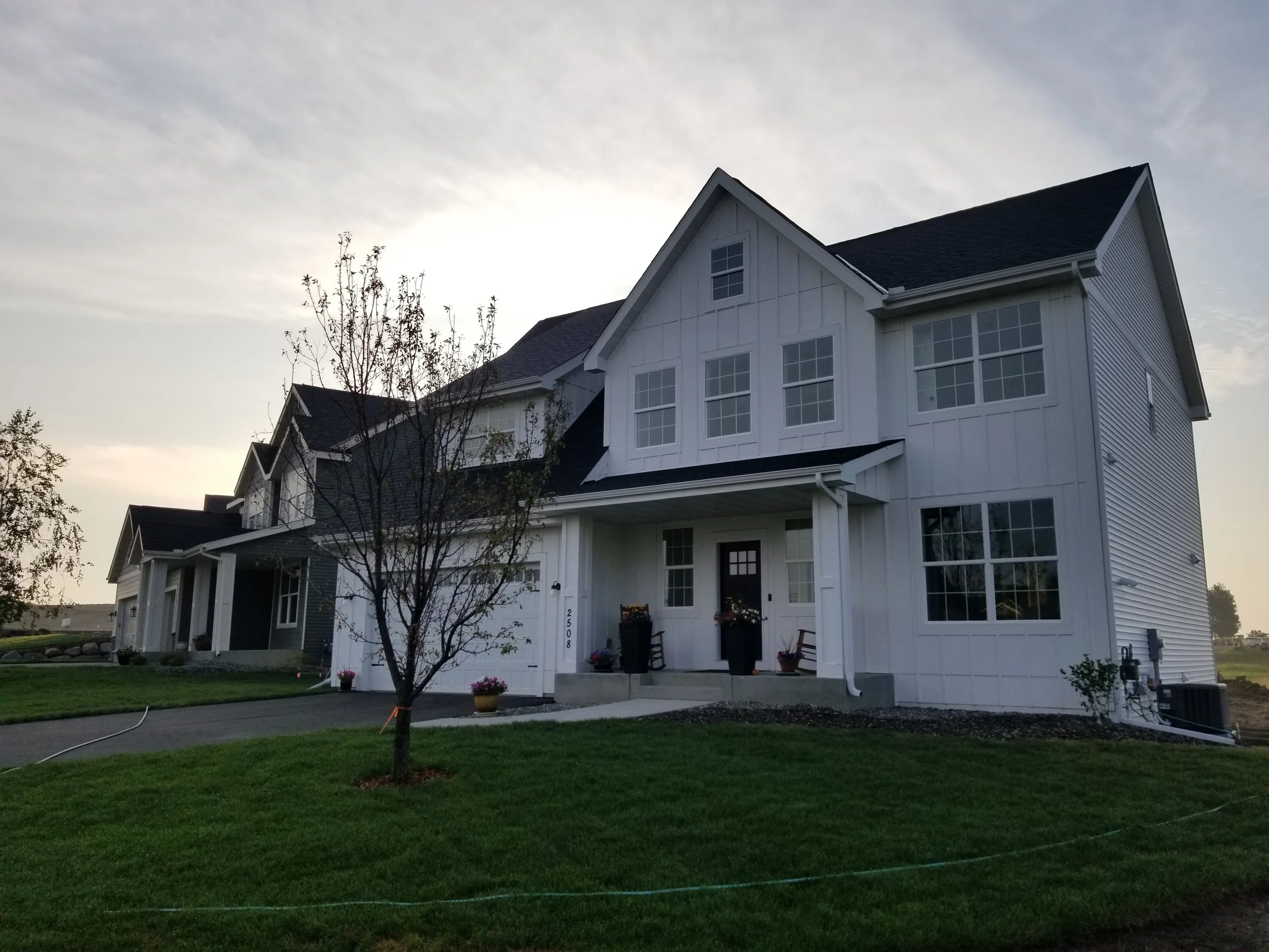 White multi-story house with large windows, a front porch with potted plants, a driveway, and a small tree in the front yard at sunset.
