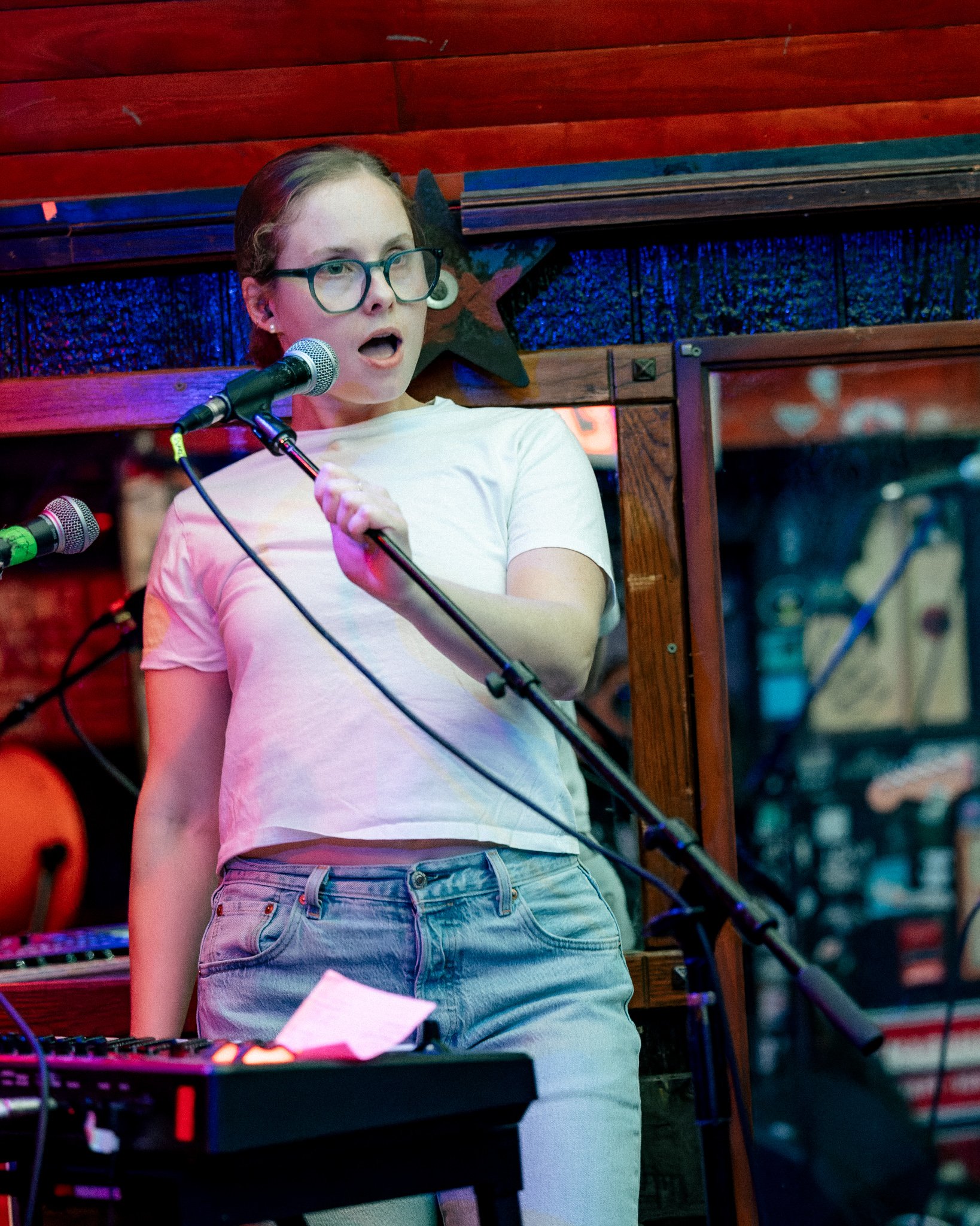 A woman with glasses and a white t-shirt singing into a microphone at a venue in Philadelphia with wooden decor and musical equipment around her.