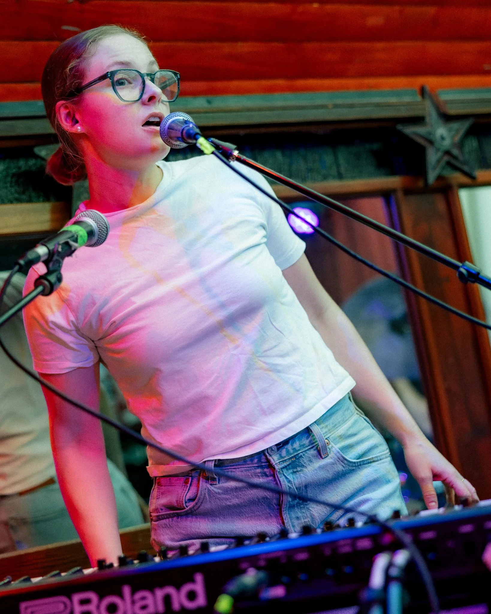 A young woman with glasses singing into a microphone on stage with colorful lighting.