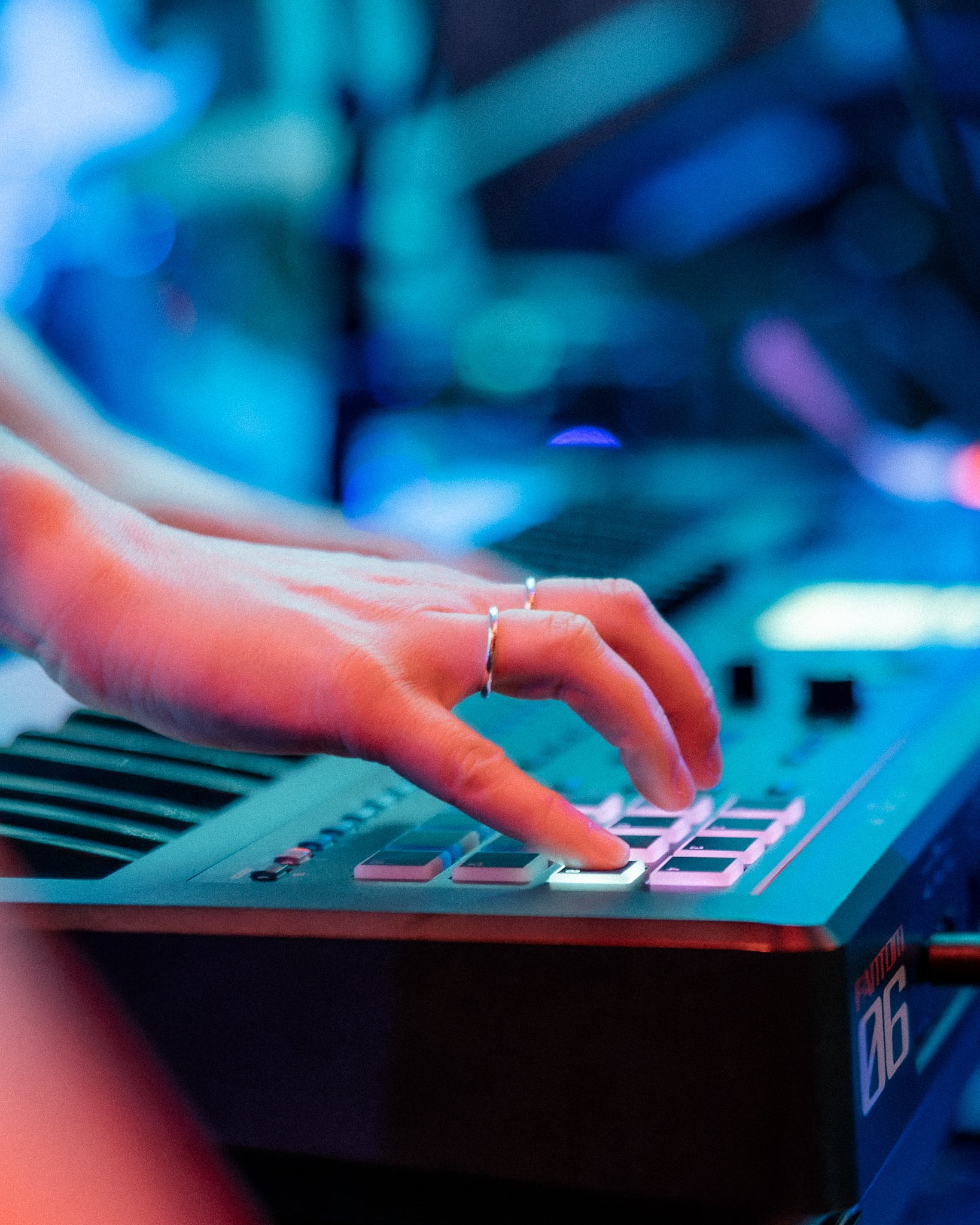 Close-up of a person's hand with rings playing a sampler on a Roland keyboard in a dimly lit venue with blue and purple lighting.