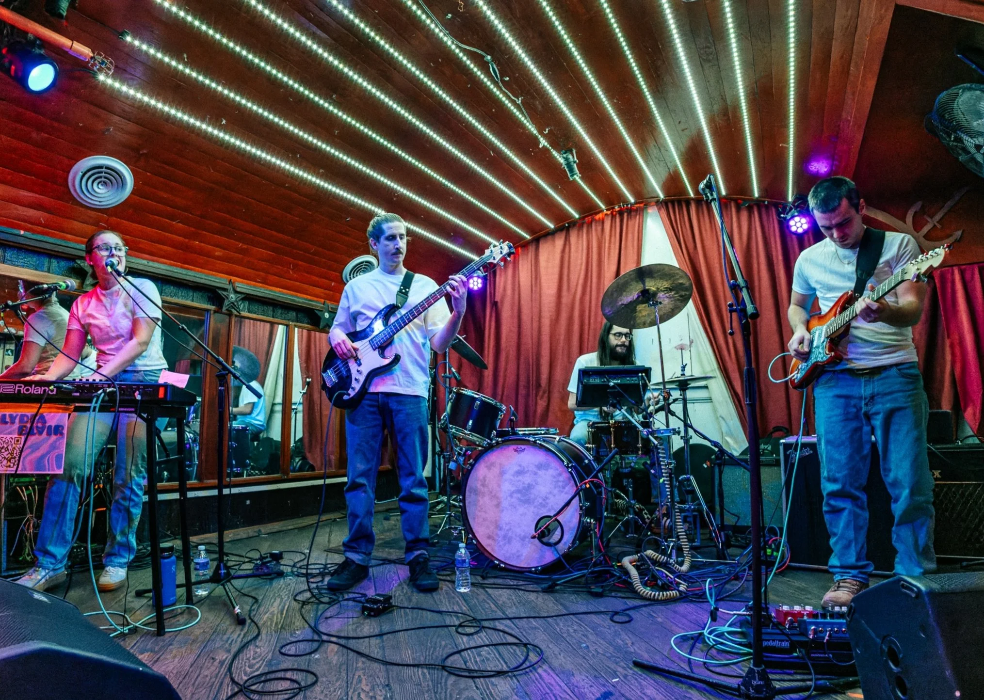 A band performing on stage with five members: a vocalist/keyboardist singing into a microphone, a bassist, a drummer, and a guitarist. The stage has colorful lighting and red curtains in the background.