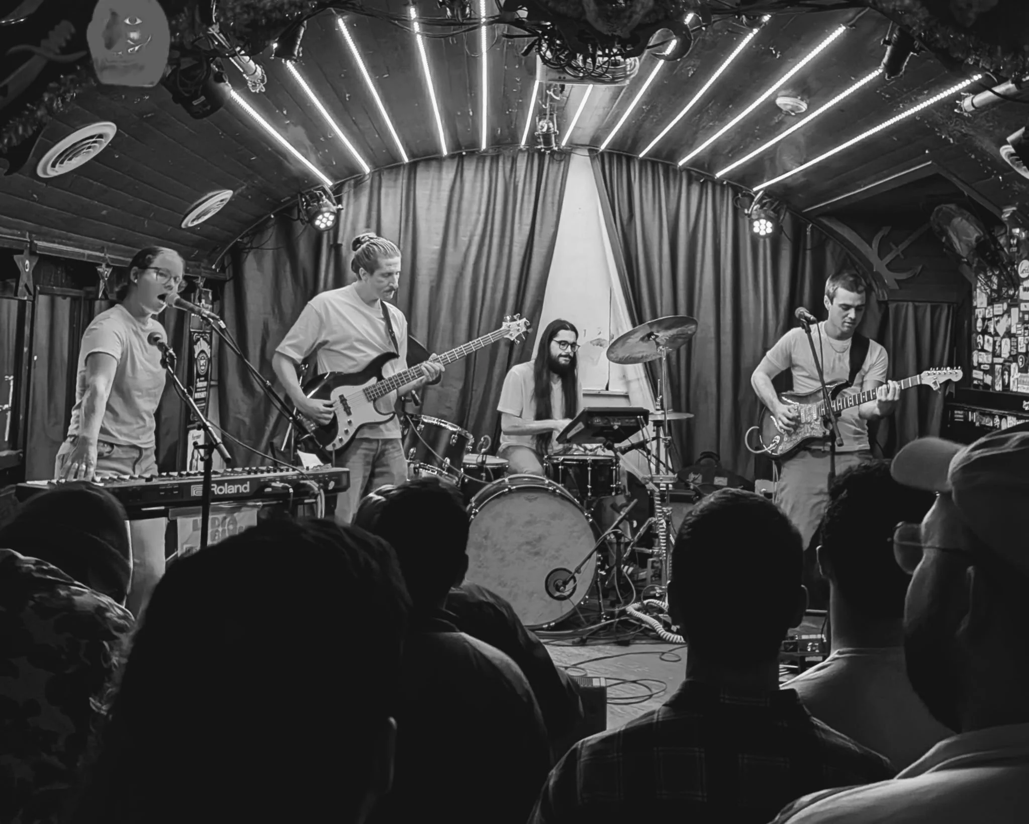 A black and white photo of the Lydia Blair band performing live on stage in front of an audience, with various musical instruments including keyboard, bass guitar, drums, and electric guitar.