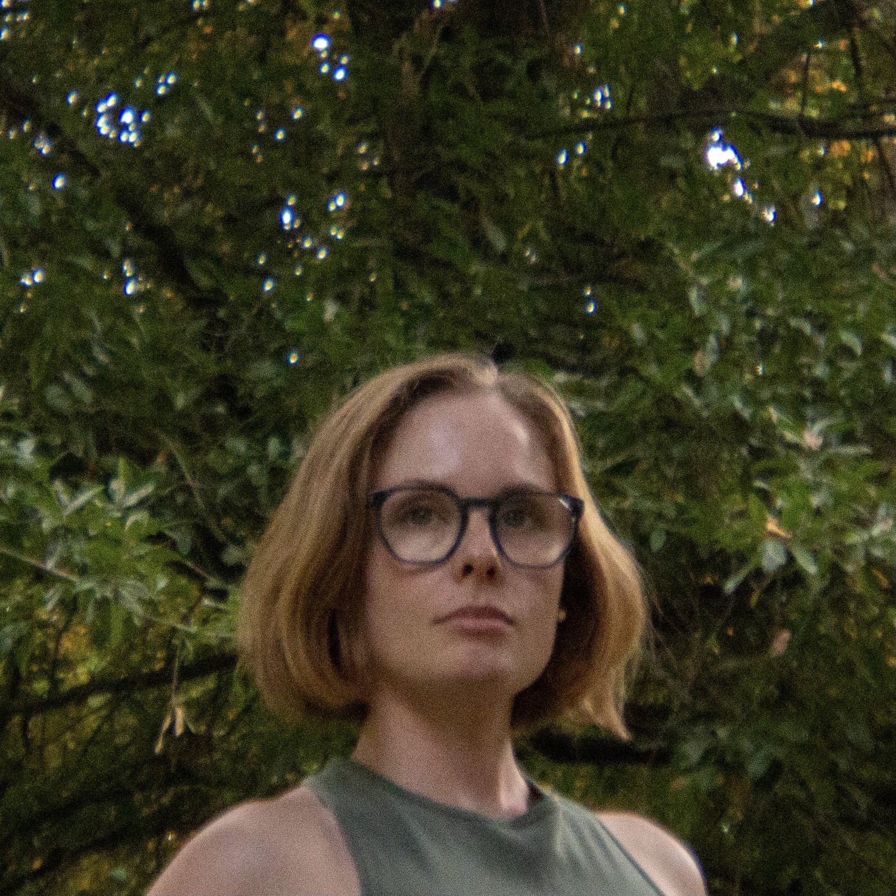 A woman with short blonde hair and navy glasses looking off into the distance outdoors with green trees in the background.