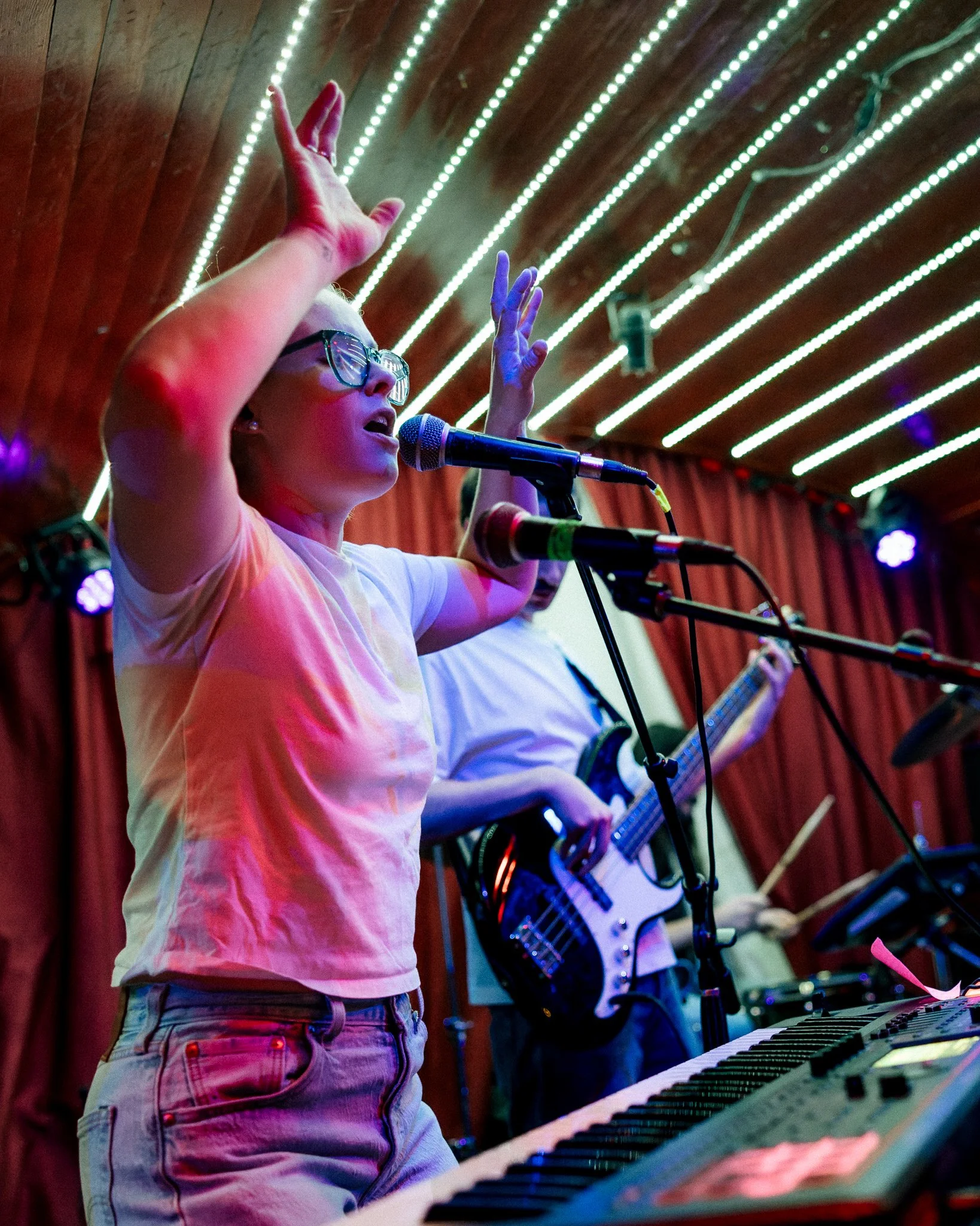A woman singing into a microphone with her arms raised during a live music performance, with other musicians playing instruments in the background, under colorful stage lighting.