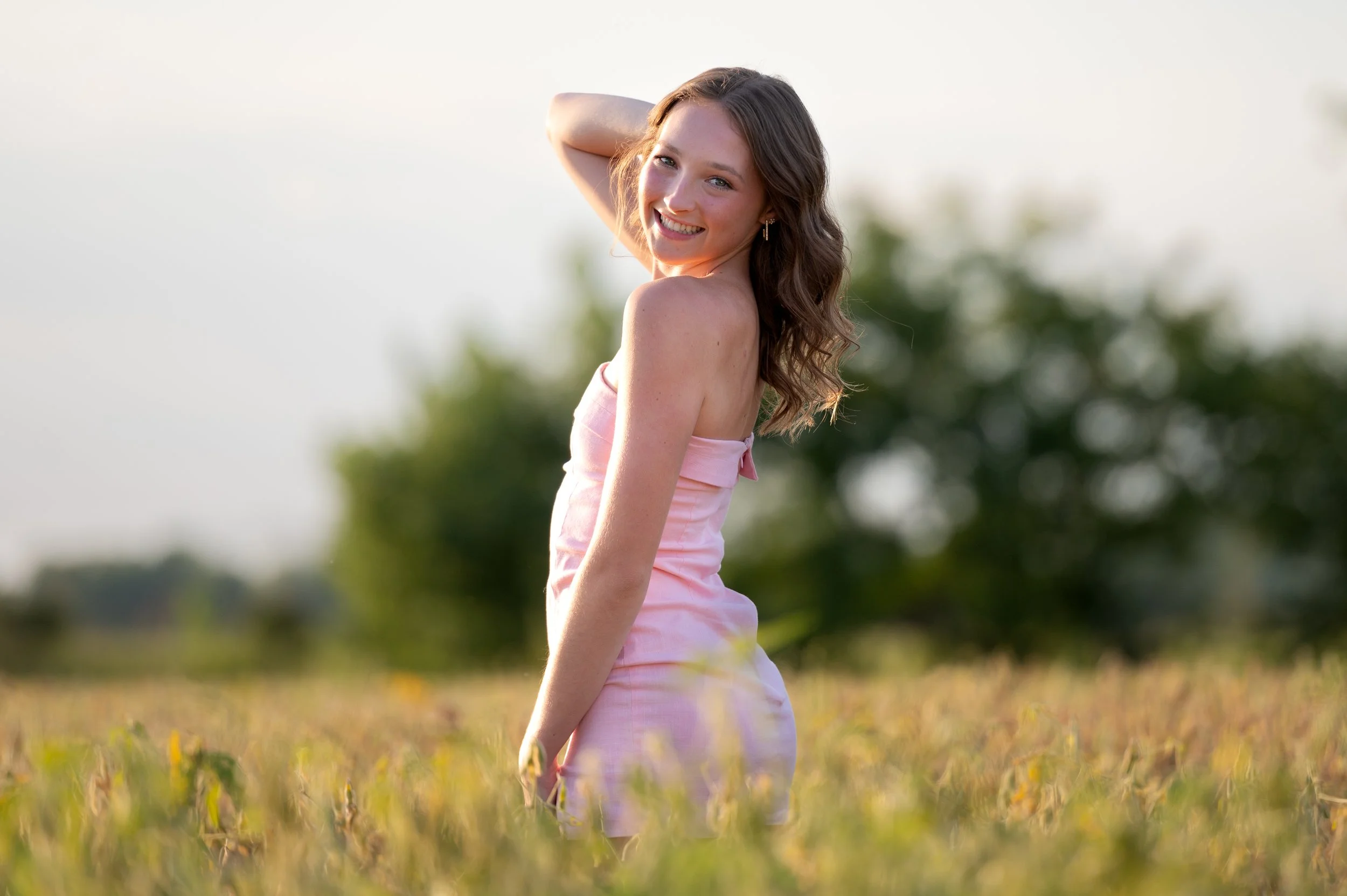 A young woman in a pink strapless dress smiling and posing outdoors in a field during daytime with blurred trees in the background.
