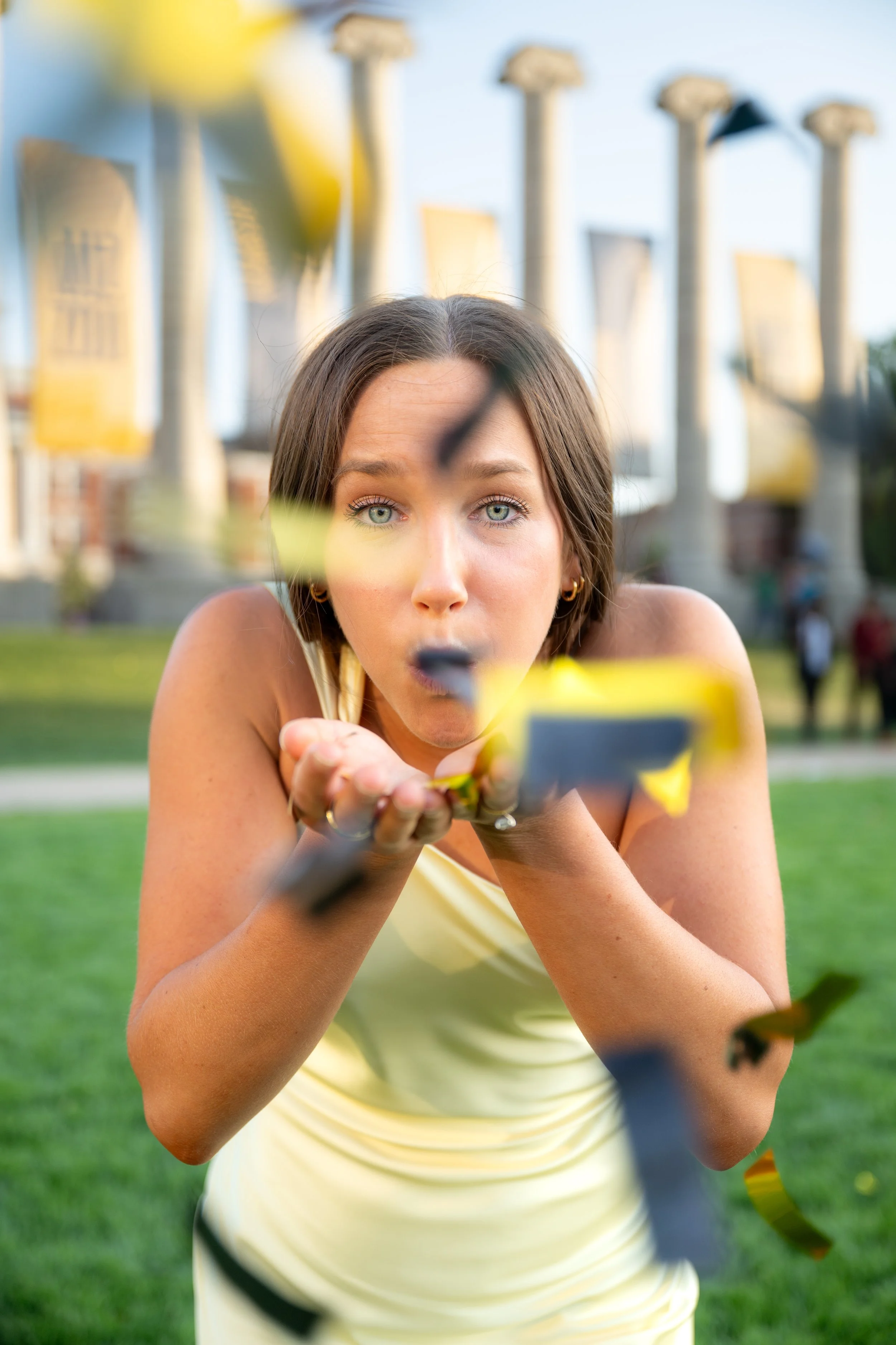 Woman in yellow dress blowing confetti through a pipe outdoors with Greek columns in the background.