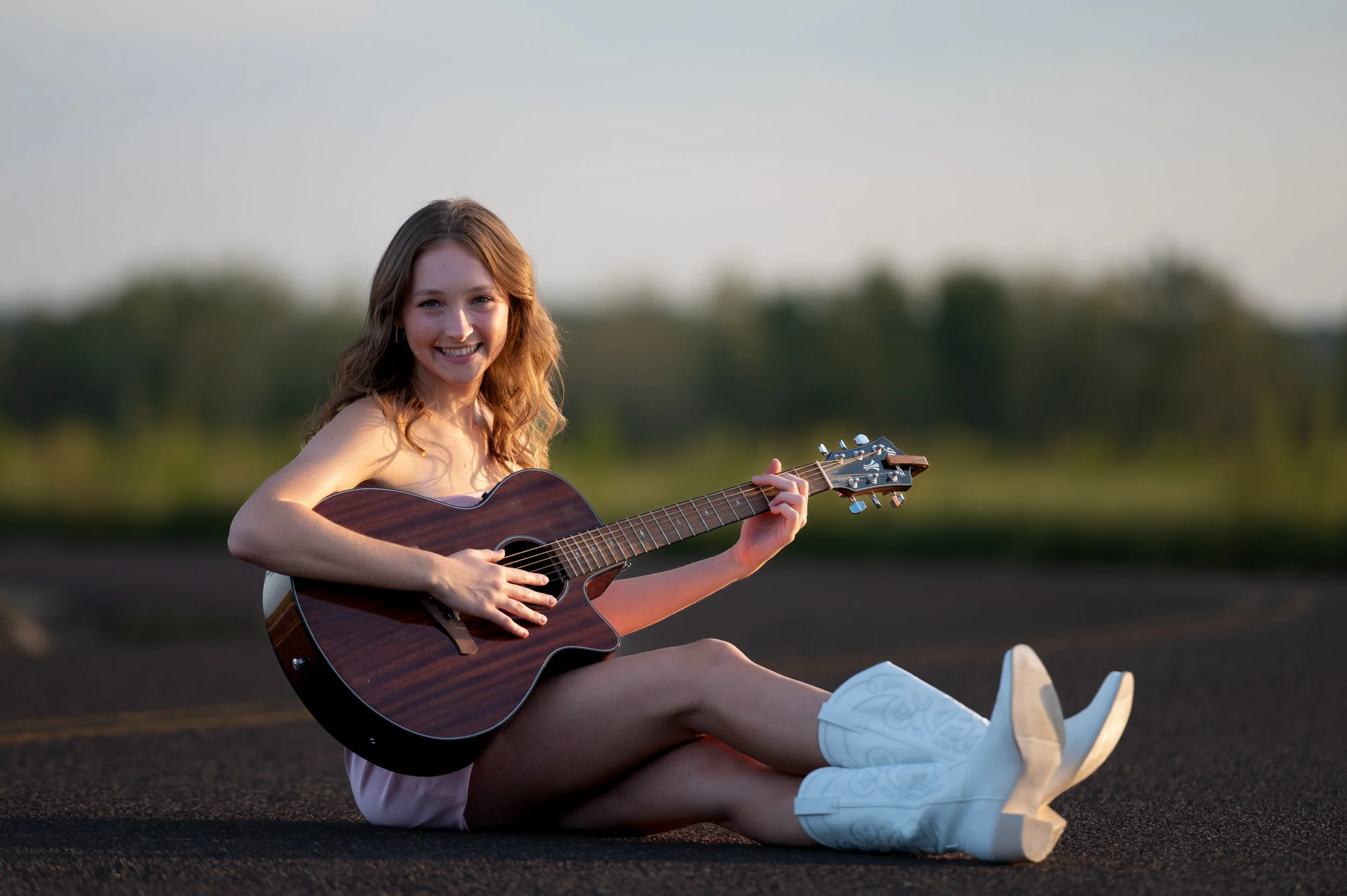 Smiling young woman sitting on the ground outdoors playing an acoustic guitar with a blurred green landscape background.
