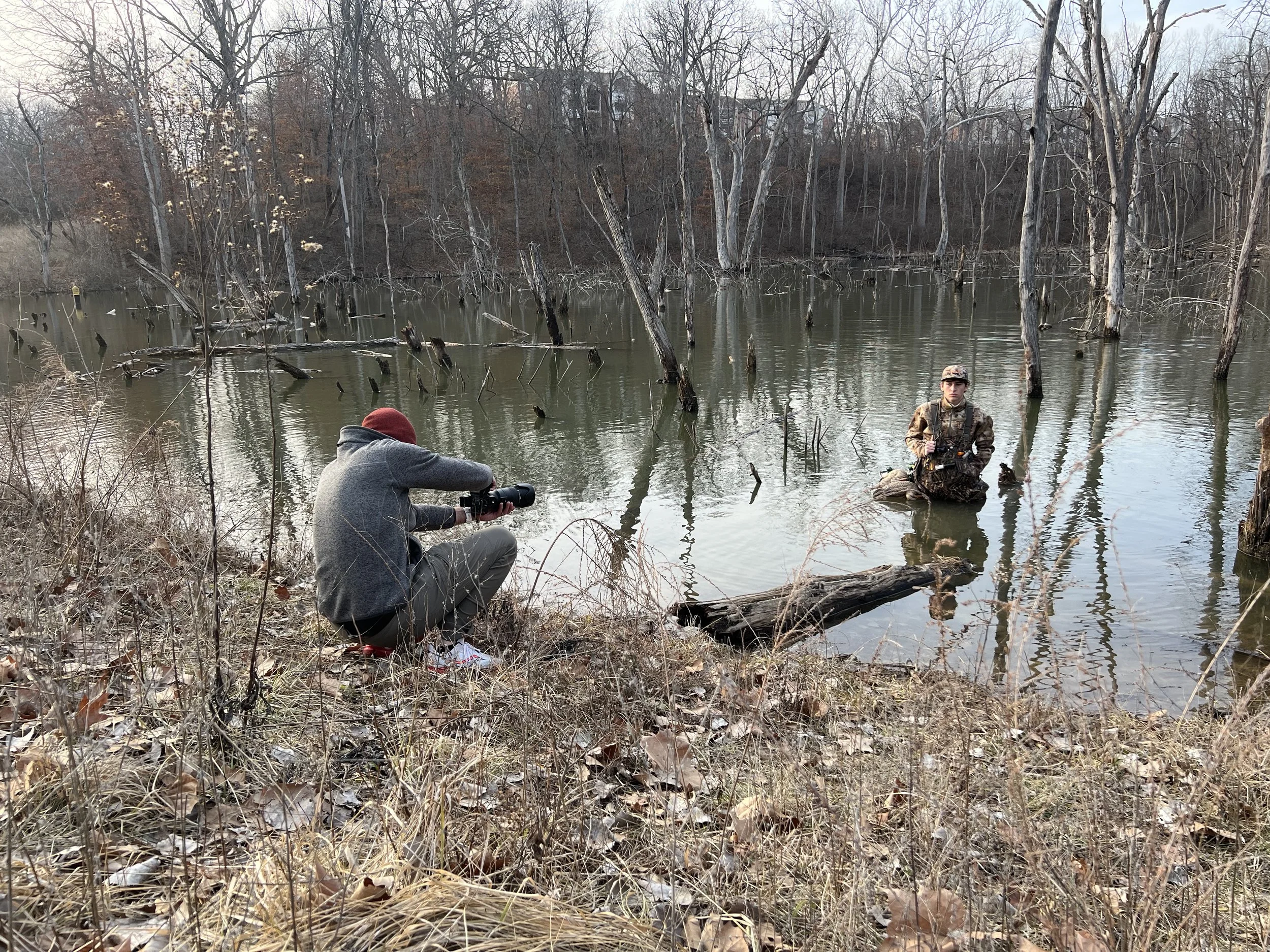 A person kneeling on the bank of a flooded area taking a photograph of a soldier standing in the water, surrounded by dead trees.