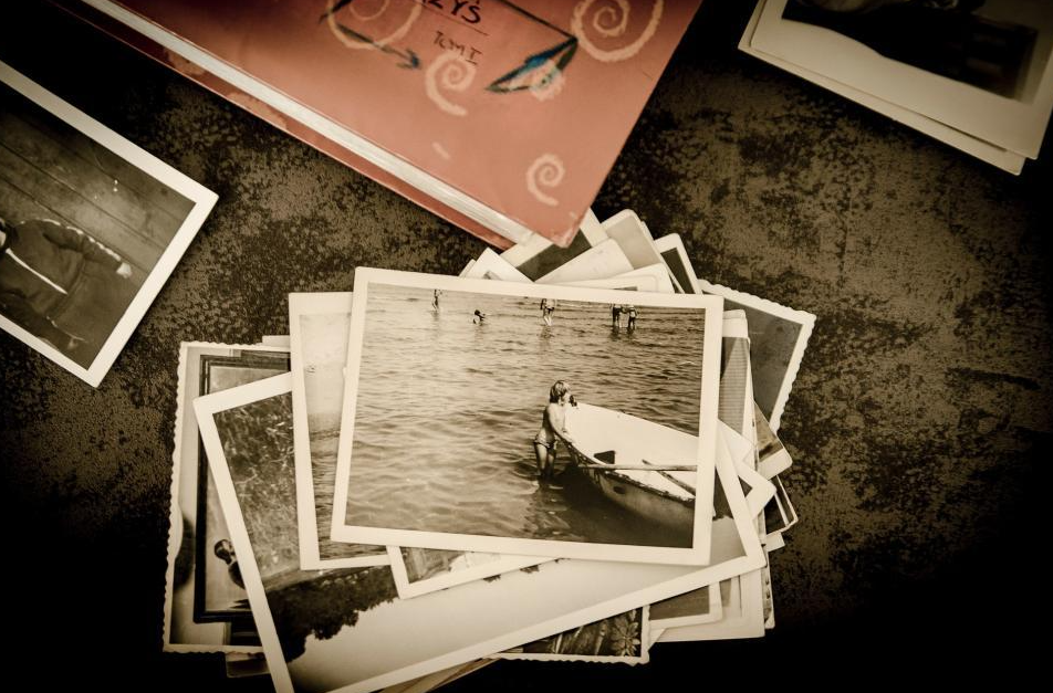 A stack of black and white photographs spread out on a dark textured surface, with one photo showing a girl by a boat in the water.