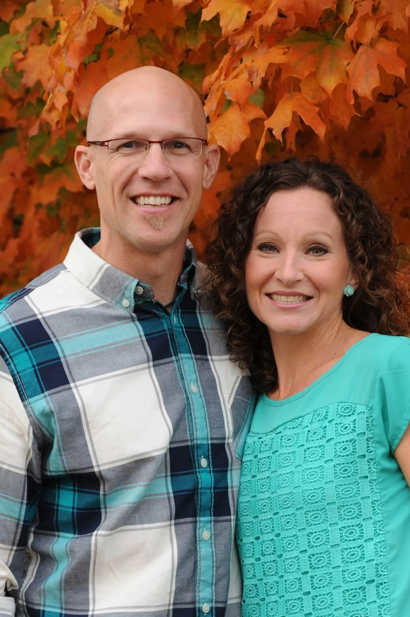 A smiling man and woman standing outdoors with autumn orange leaves in the background.
