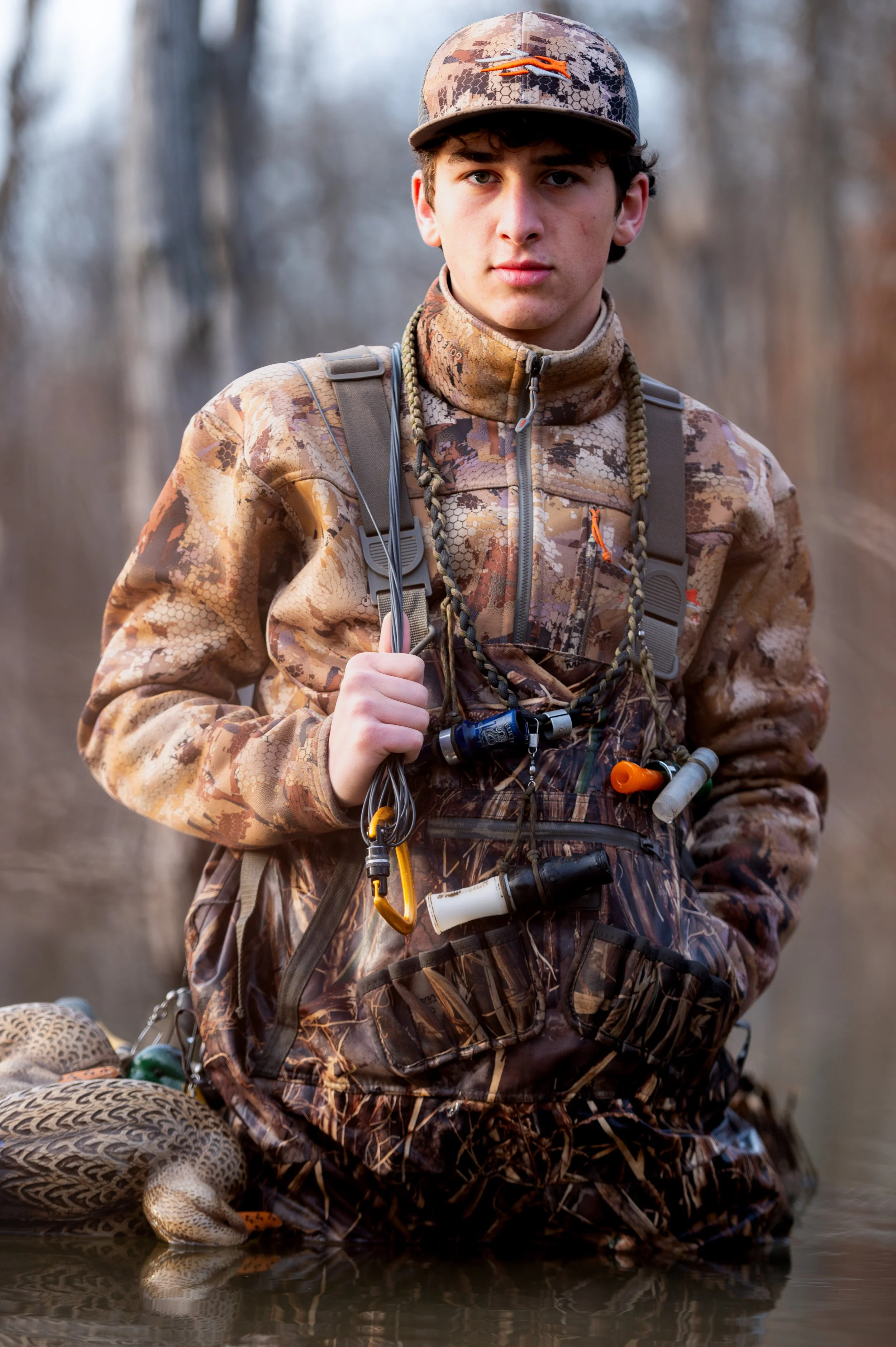 Young man in camouflage outdoor gear standing in water during a hunt, holding a hunting gear with a duck near his leg, in a wooded area.