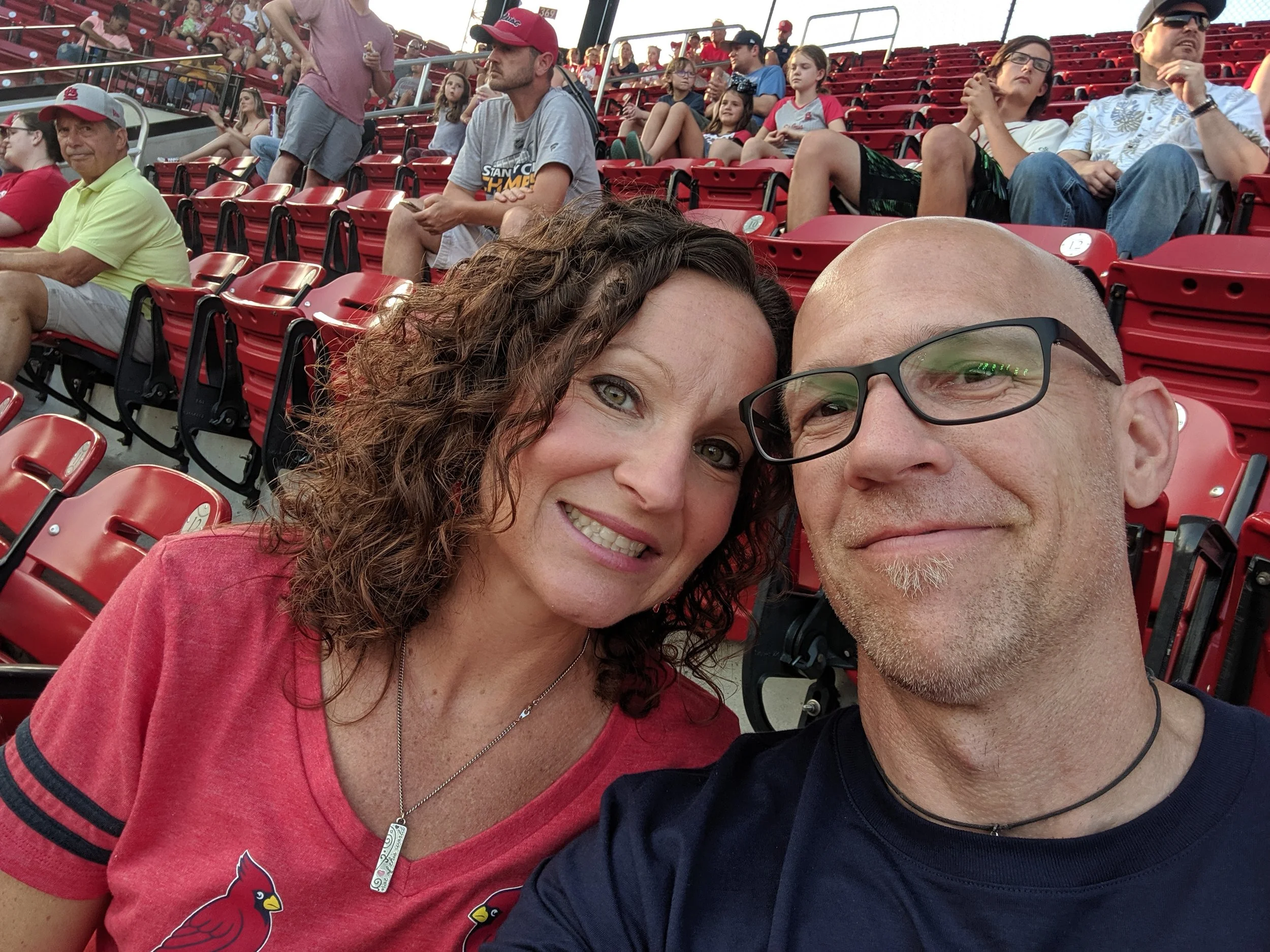 A woman and a man taking a selfie at a baseball game, seated in a stadium with red seats. Other spectators are visible in the background.