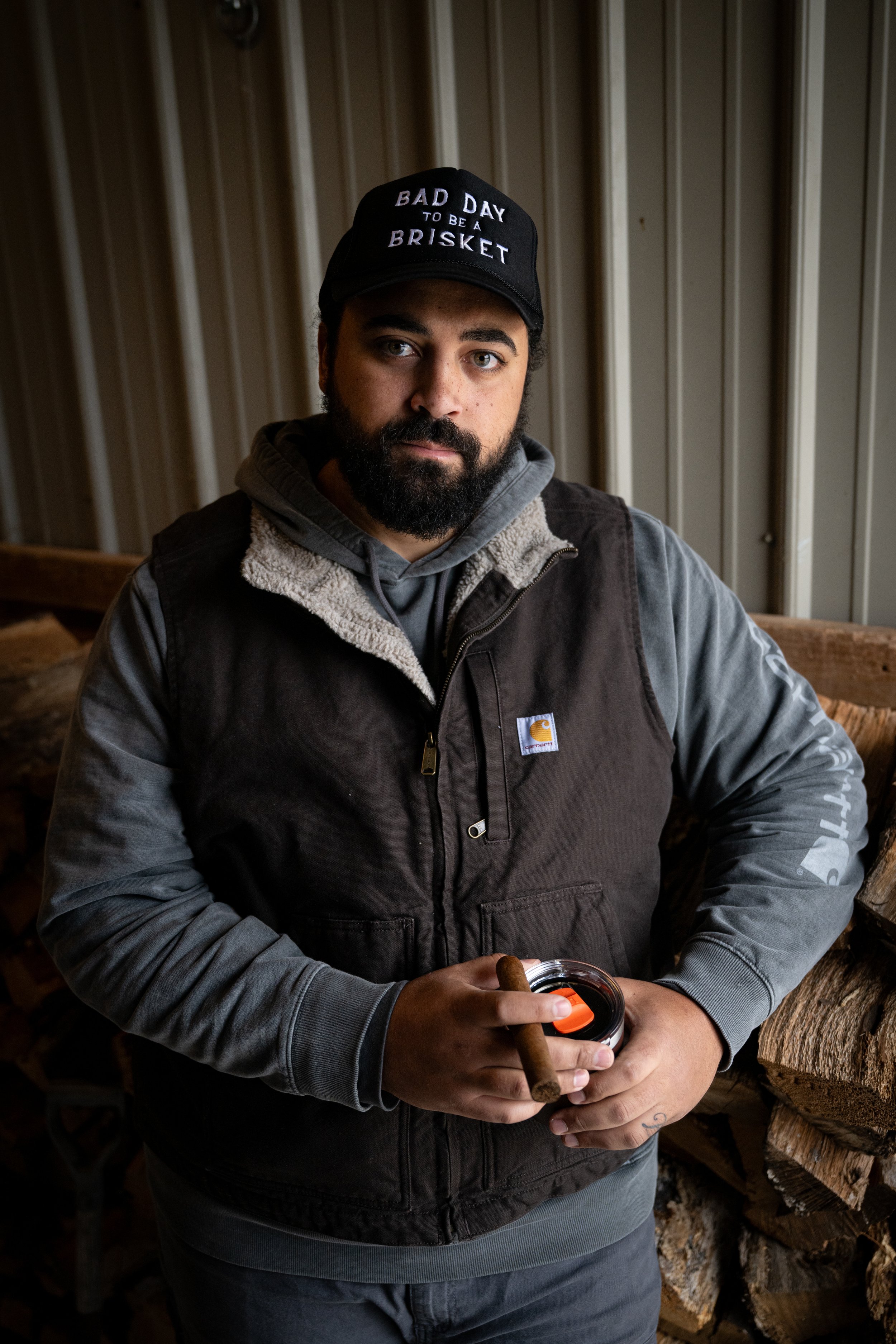 A man with a beard wearing a black cap that says 'Bad Day to be a Brisket', a layered jacket, and a gray hoodie, holding a cigar and a beverage container, standing indoors in front of a woodpile and metal wall.