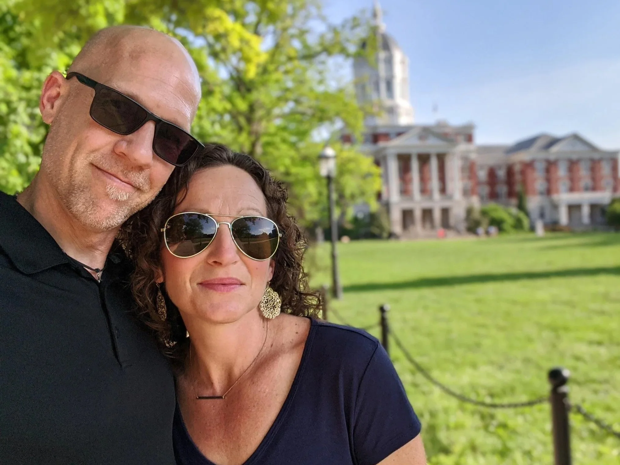 A man and woman wearing sunglasses outdoors with a historic brick building and green trees in the background.