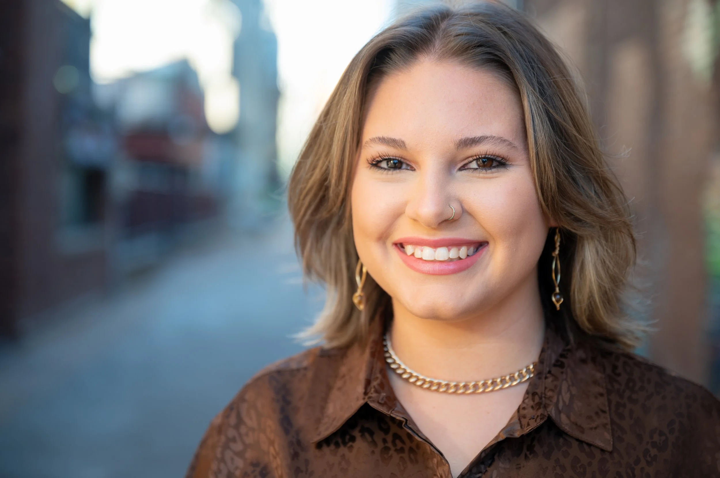 Close-up of a young woman with shoulder-length light brown hair, wearing a brown patterned blouse, gold jewelry, and a nose ring, smiling outdoors in front of a blurred city background.