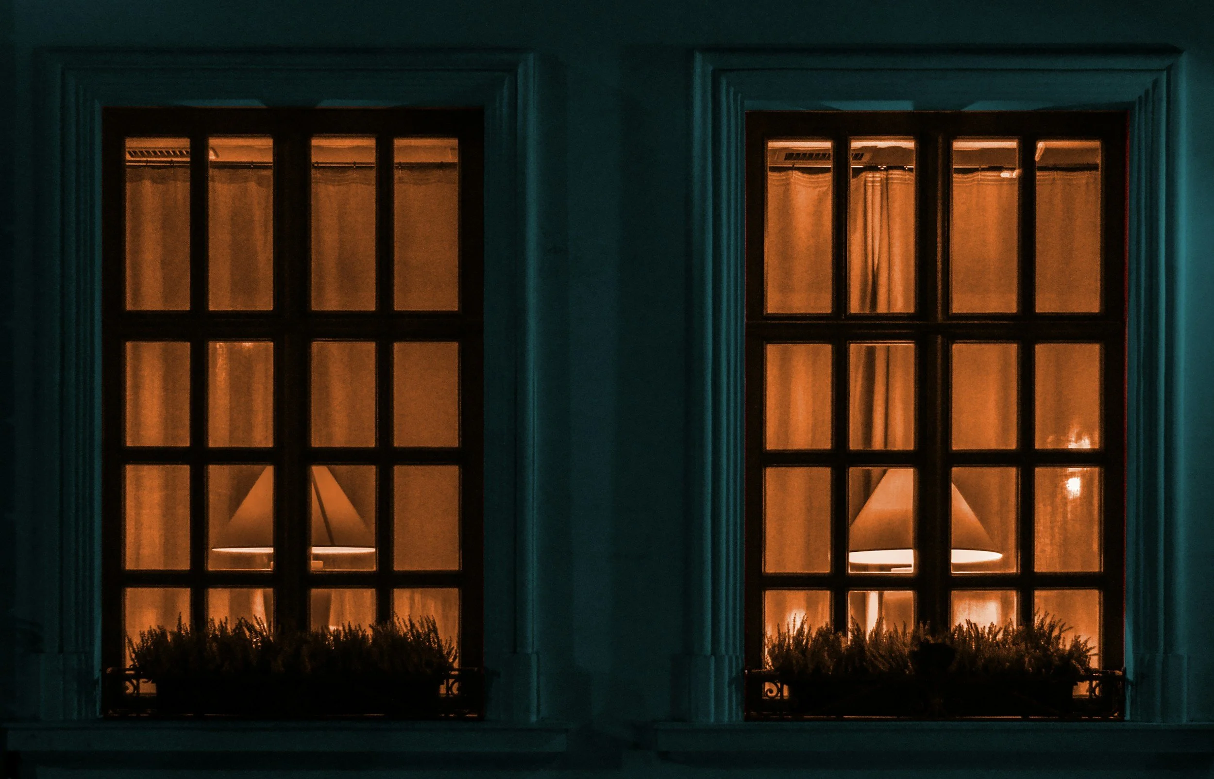 Two illuminated windows with curtains and lamps visible inside, lit warmly, against a dark exterior wall.