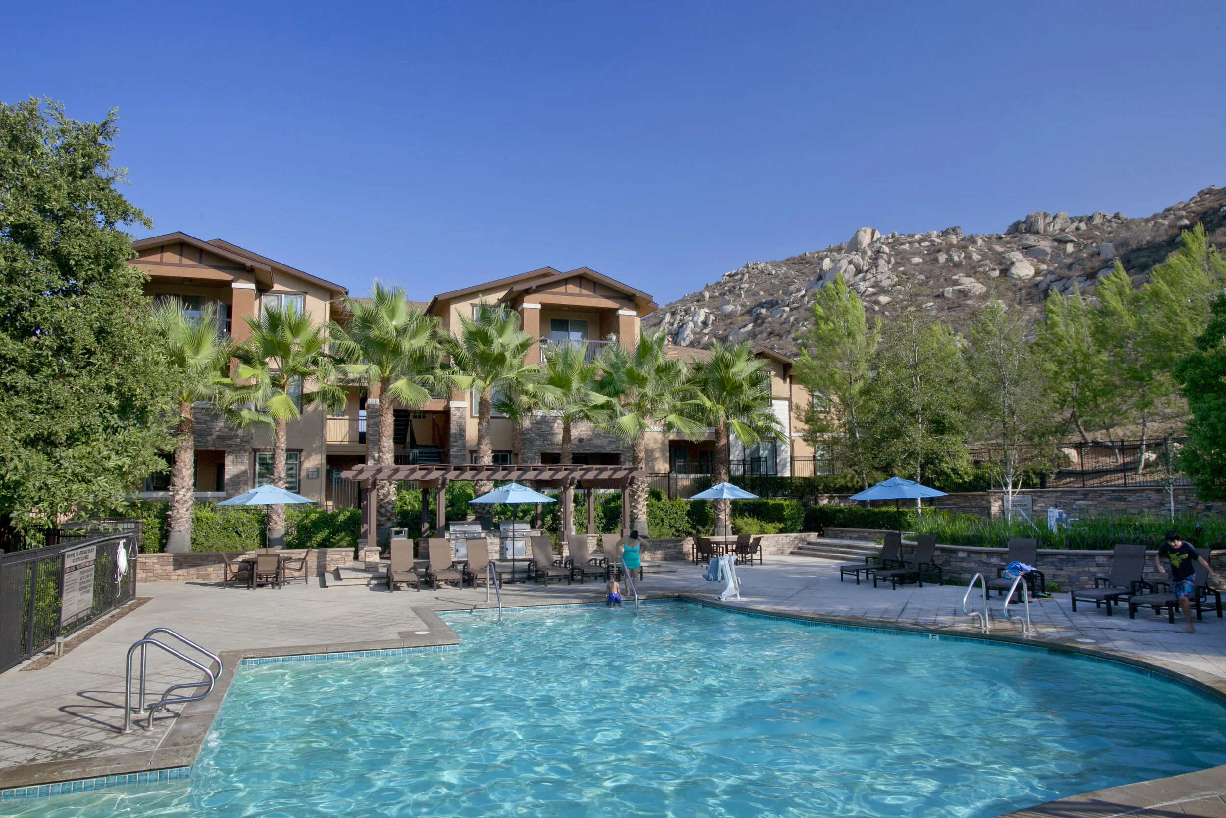 Sparkling pool at The Overlook at Rancho Belago in enclosed pool yard surrounded by lounge chairs, tables shaded by umbrellas, lush greenery, and mountain views.