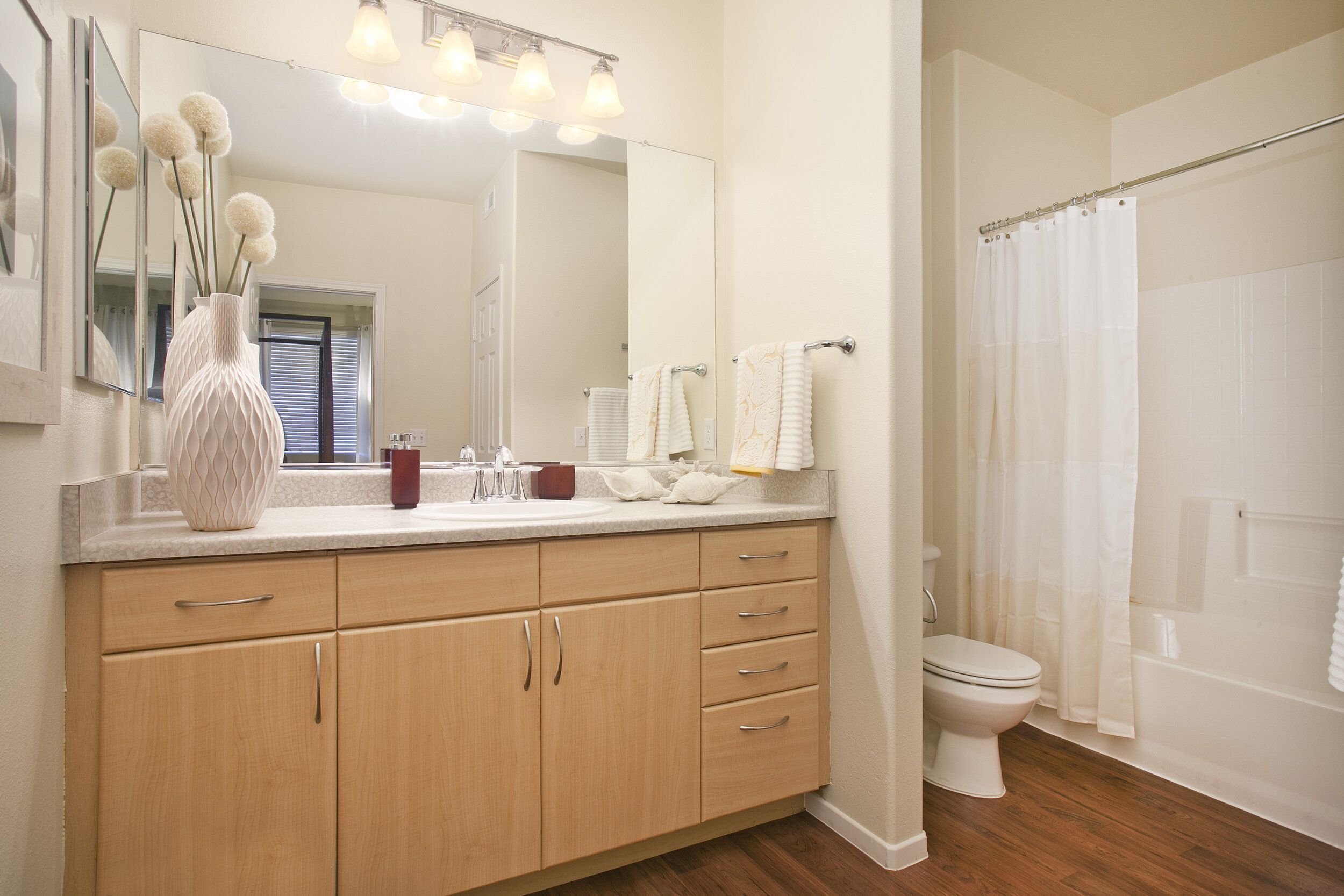 Bathroom in apartment at The Overlook at Rancho Belago with wood plank flooring, light wood cabinets, shower/tub, and large mirror.