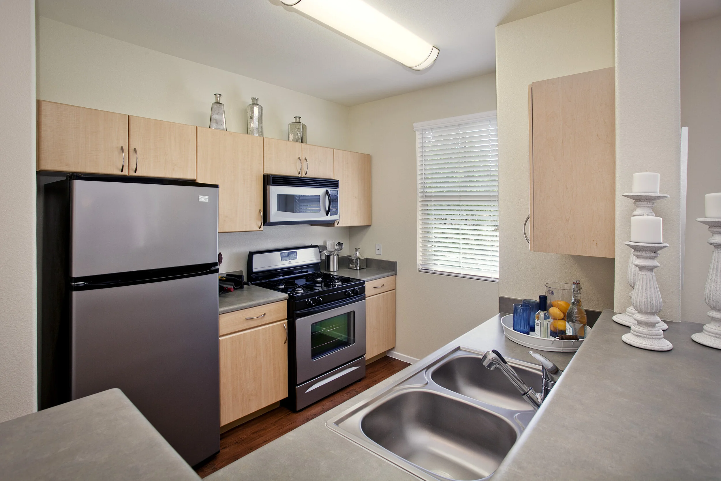 Kitchen in apartment at The Overlook at Rancho Belago with light wood cabinets, stainless steel appliances, wood plank flooring, cream walls, and window. 