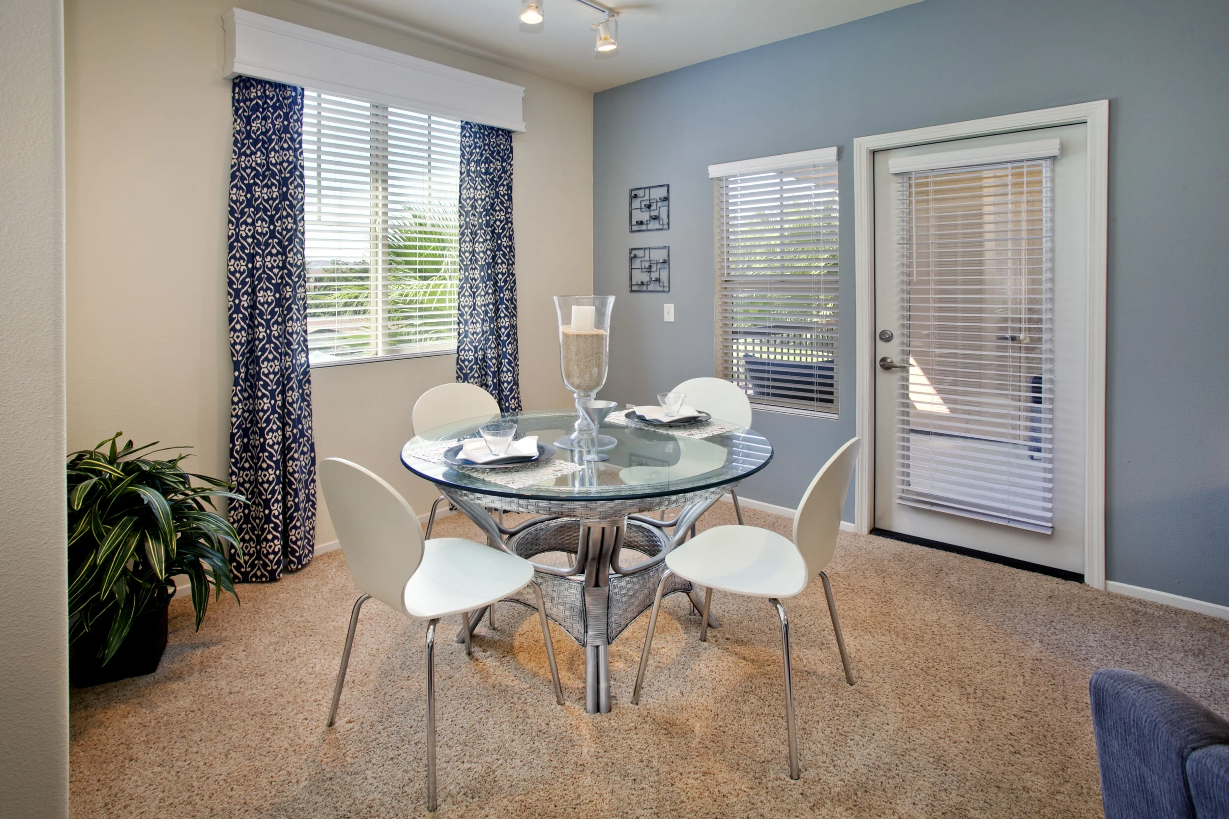 Dining area inside apartment at The Overlook at Rancho Belago with carpeted flooring, cream walls, one blueish grey accent wall, and large windows. 