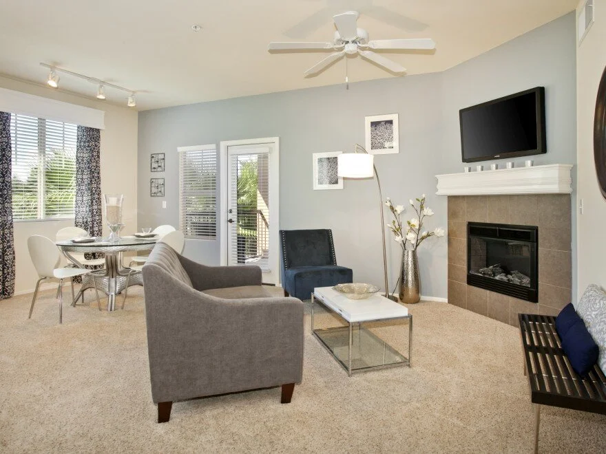 Living/dining area at The Overlook at Rancho Belago with carpeted flooring, cream walls, one blueish grey accent wall, fireplace, and large windows. 