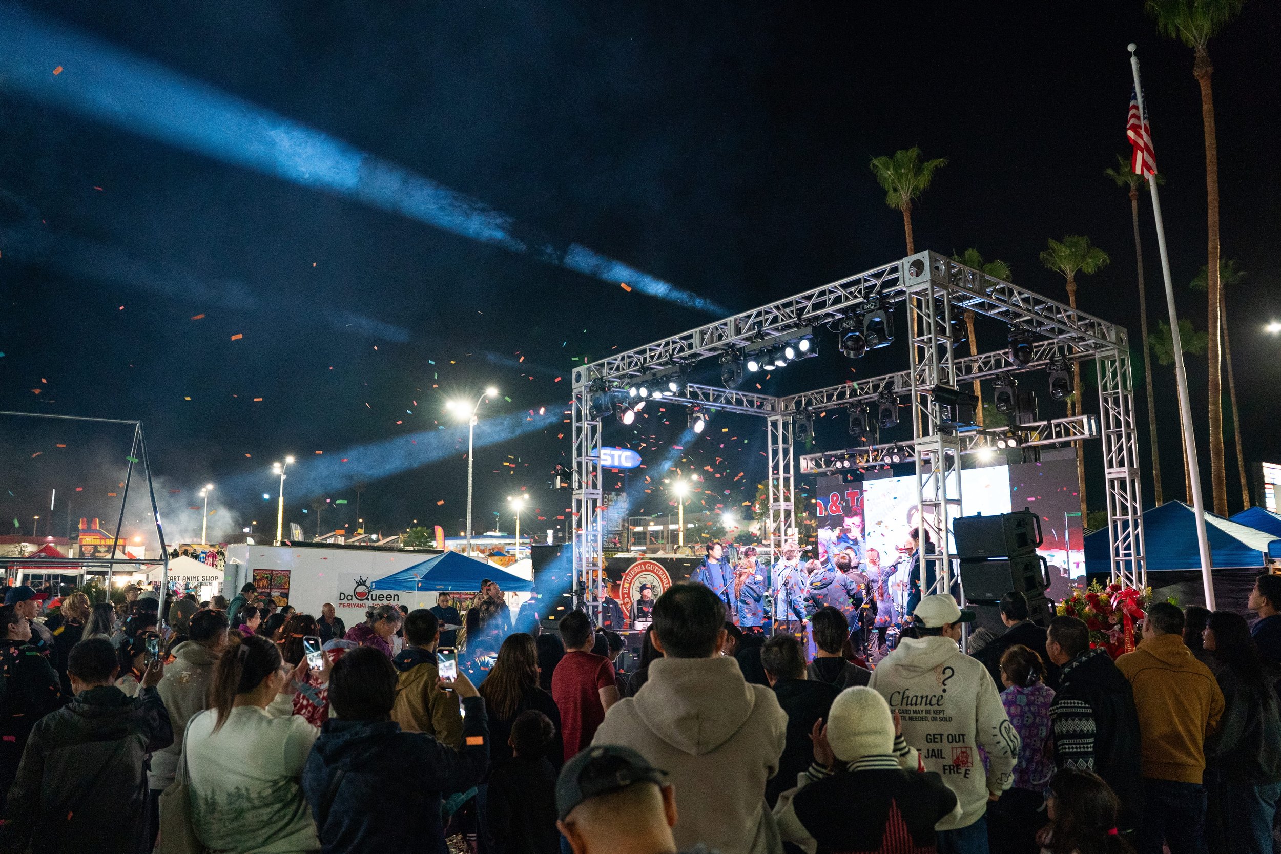 A stage with performers and a large crowd gathered underneath. There are lights and confetti in the sky. 