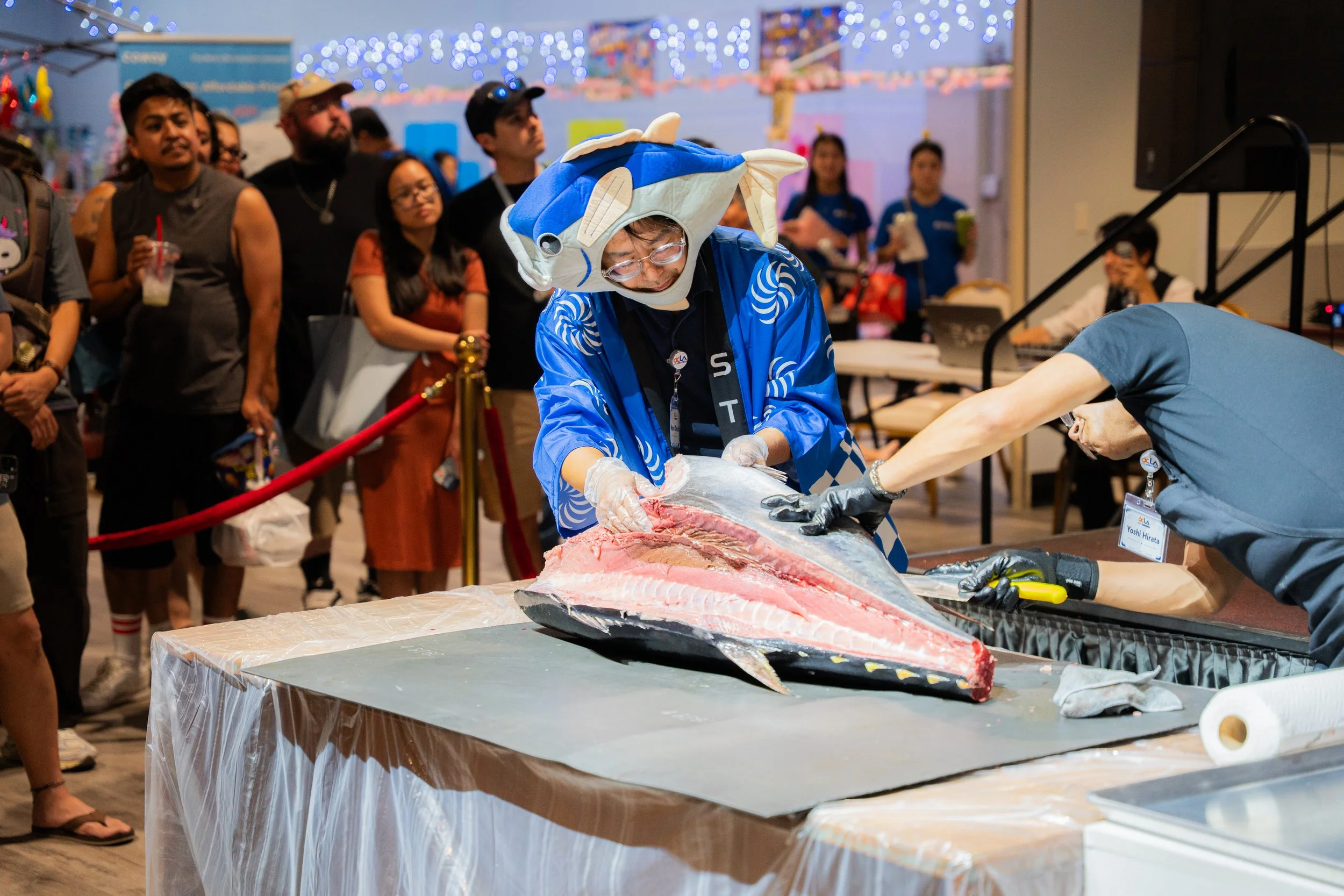 A large crowd at the Legacy Pavilion at the STC Rowland Legacy Natsu Festival watching the Bluefin Tuna Cutting Show. A man wearing a fish costume and traditional Japanese robe helps handle the blue fin tuna as the sushi chef demonstrates the cutting