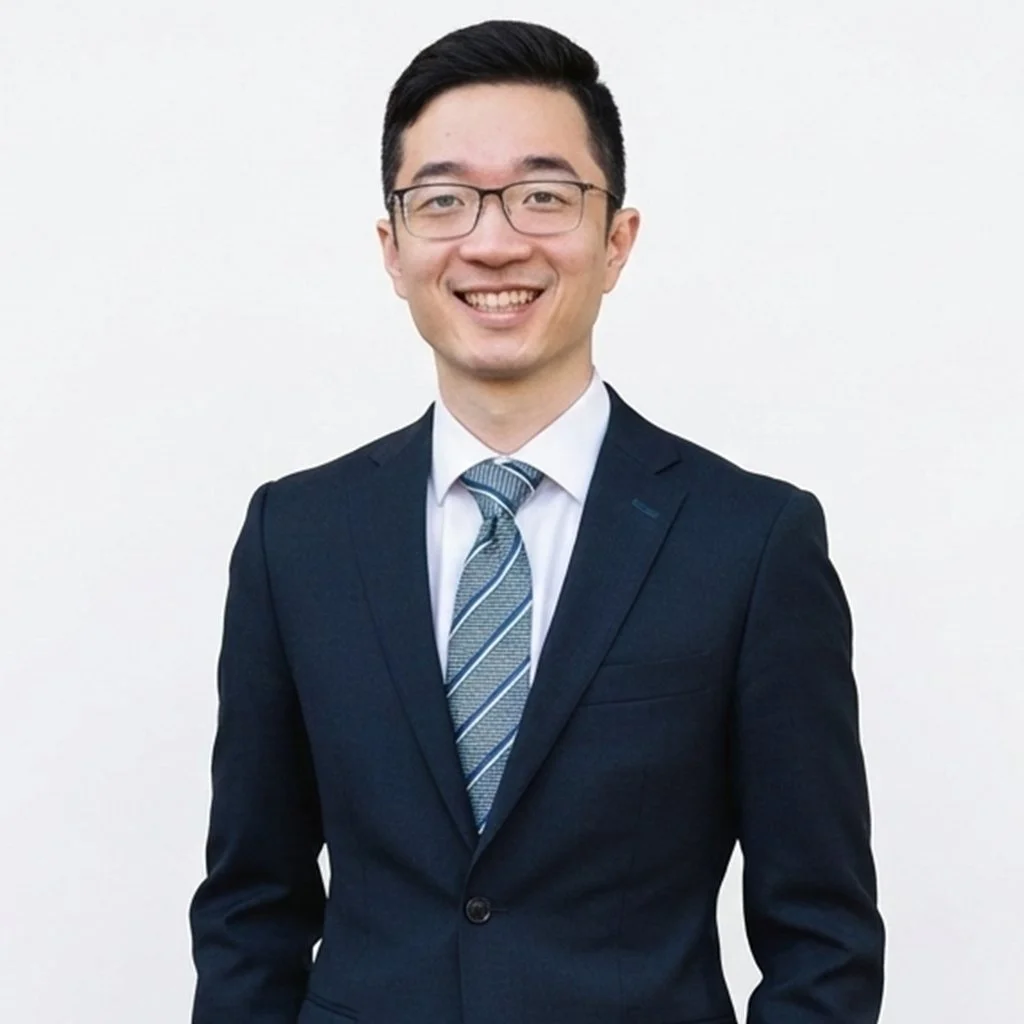 A young man with glasses in a business suit, smiling, standing against a plain white background.